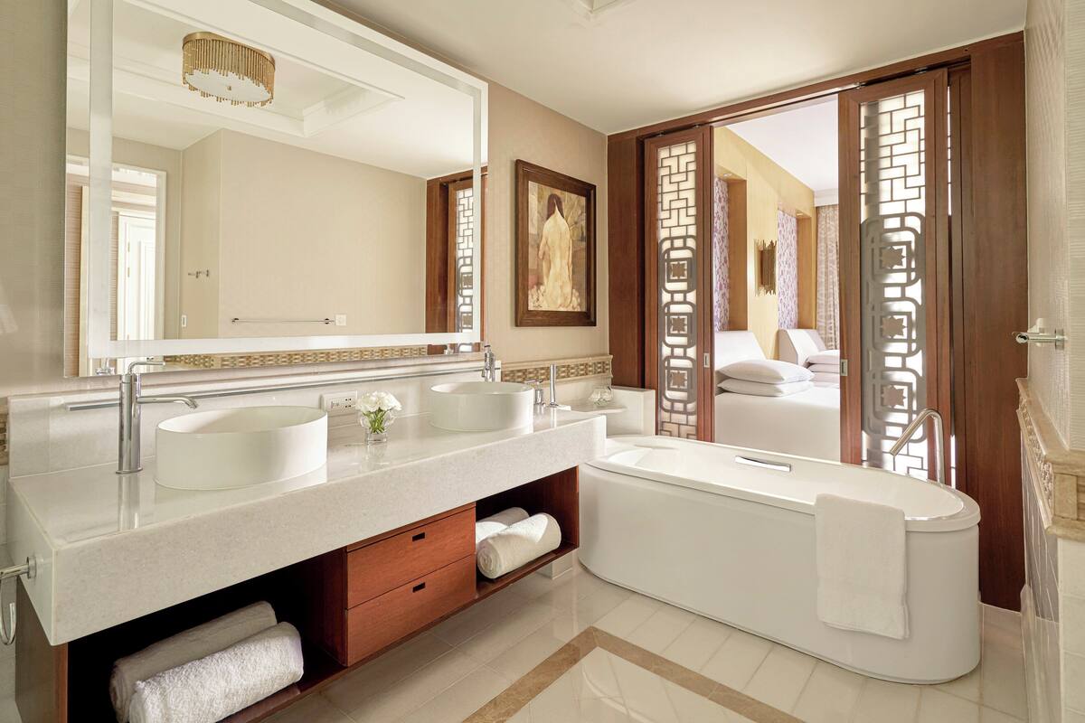 A modern bathroom with a large mirror, two sinks, a bathtub, and a wooden door with a decorative screen.