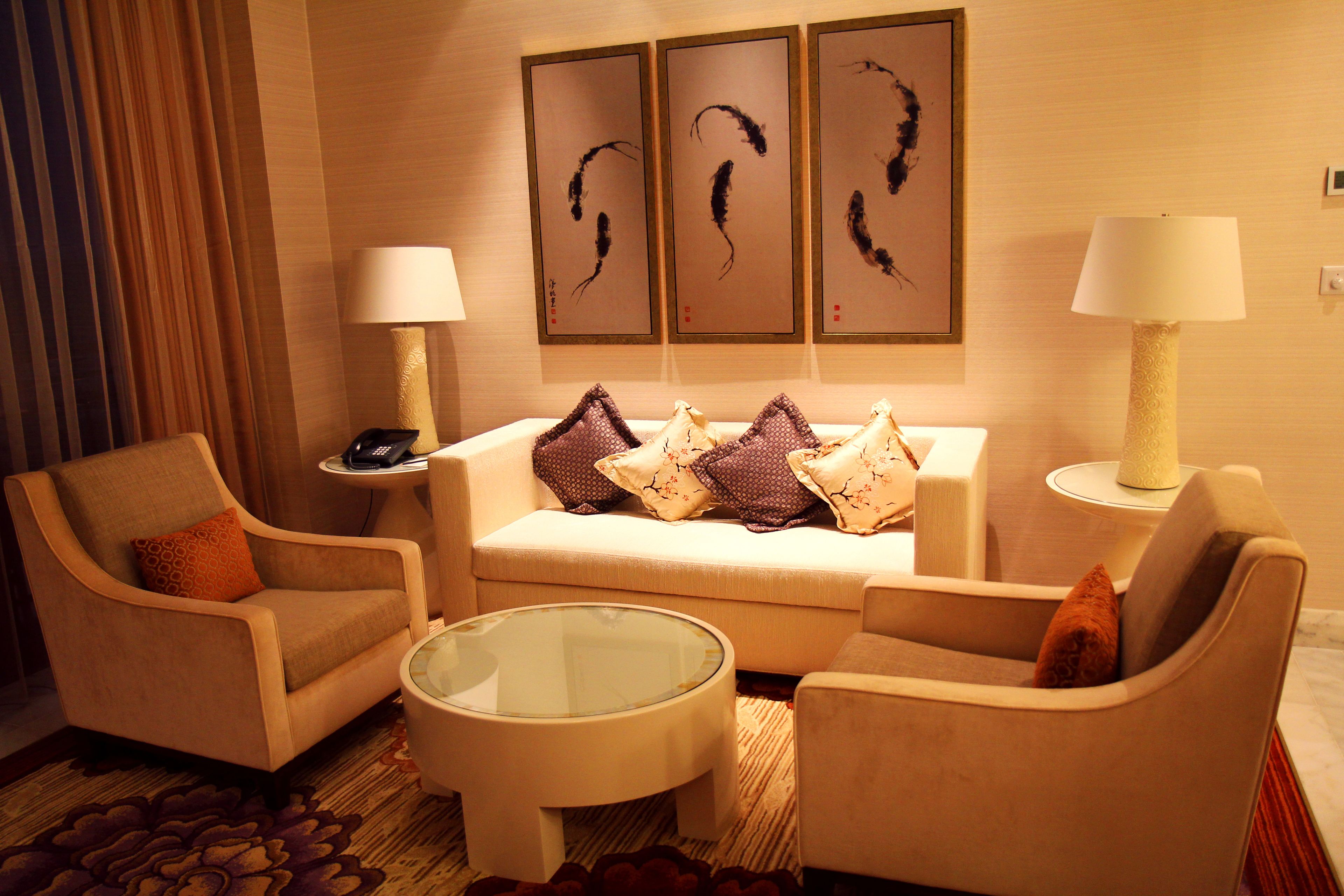 Een hotelkamer met een bank, twee fauteuils, een glazen salontafel en een bijzettafeltje met een lamp. Boven de bank hangen drie kunstwerken in lijsten.