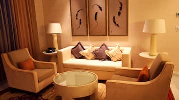 Een hotelkamer met een bank, twee fauteuils, een glazen salontafel en een bijzettafeltje met een lamp. Boven de bank hangen drie kunstwerken in lijsten.