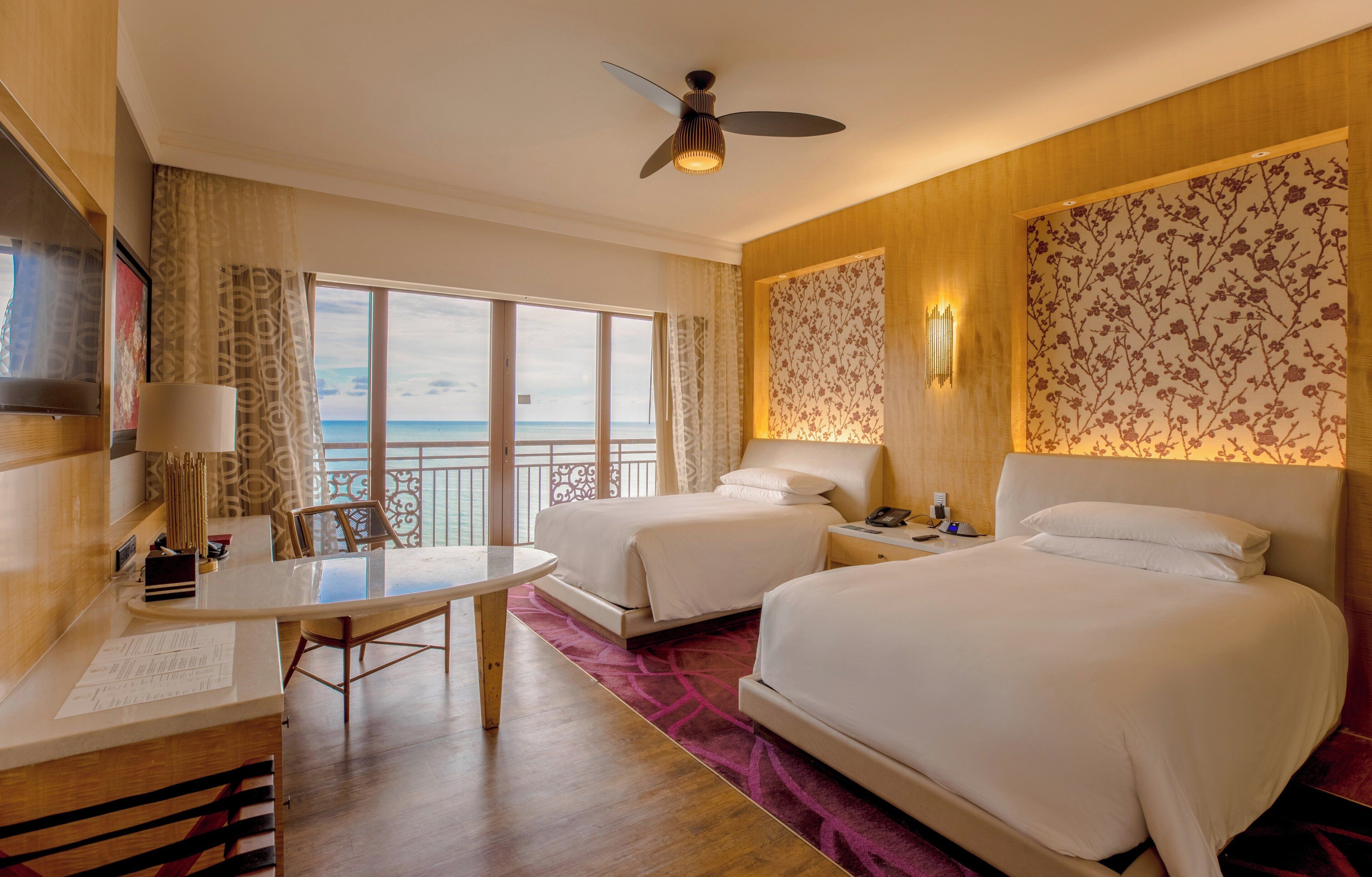 A hotel room with two beds, a desk, a chair, a table, a ceiling fan, and a view of the ocean.