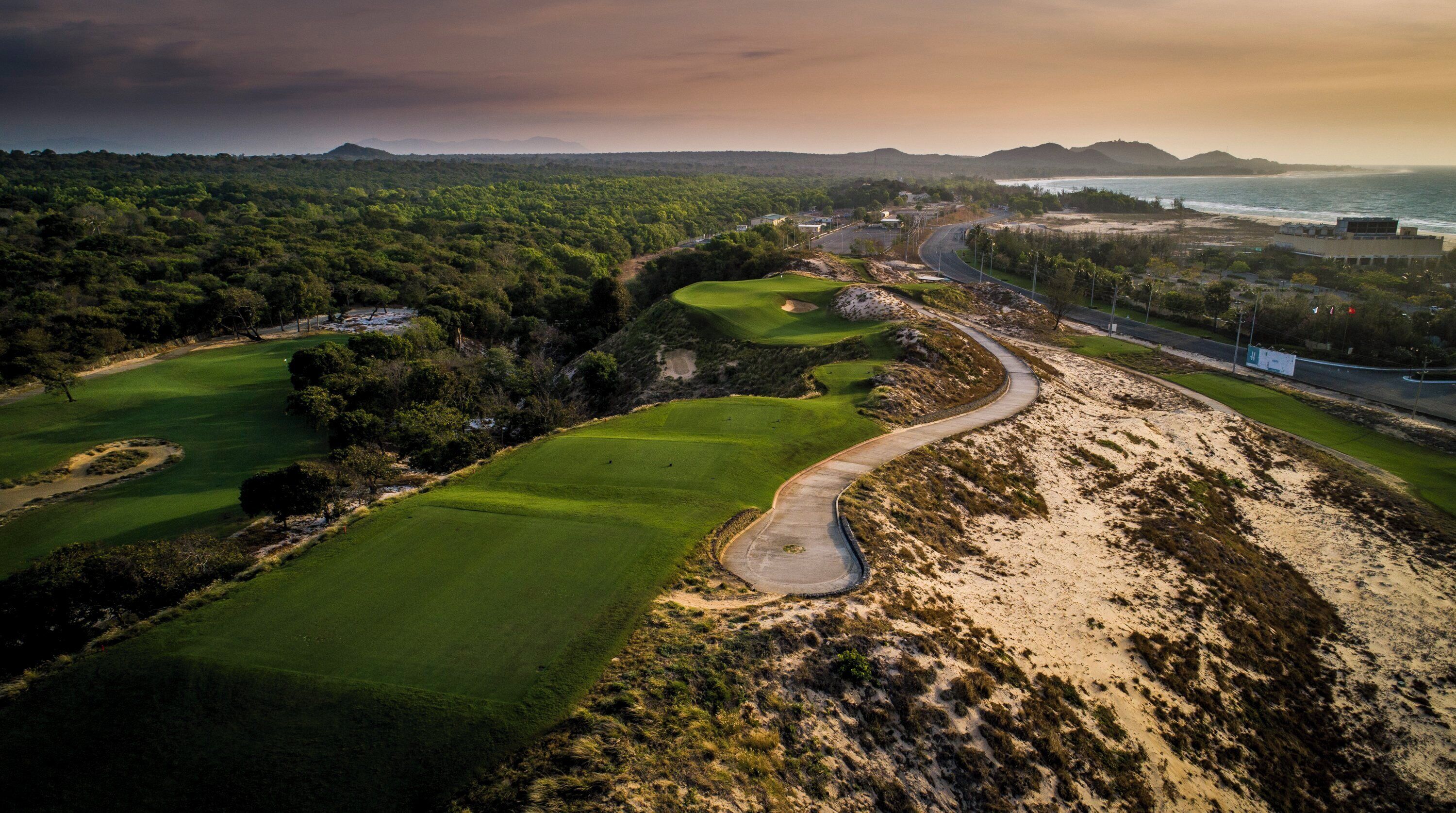 Vue aérienne d’un terrain de golf avec des bunkers de sable, une route sinueuse et un paysage côtier en arrière-plan.