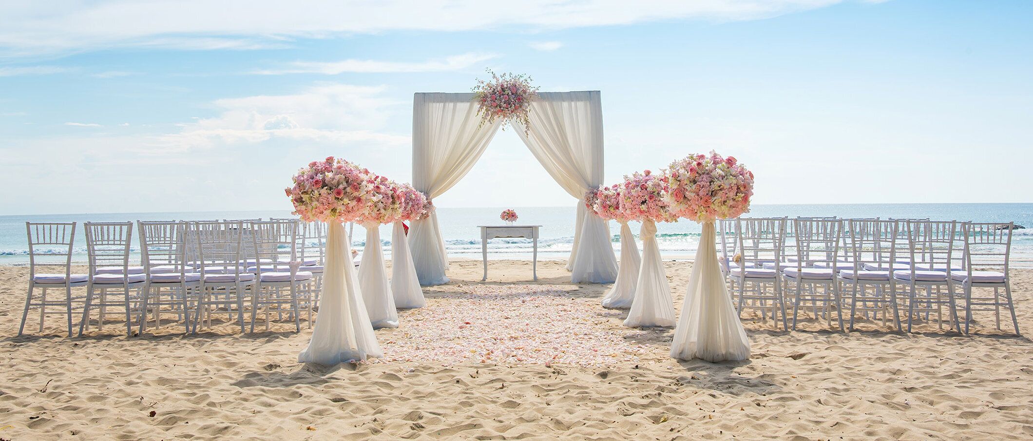 En bröllopsdekoration på stranden med vita stolar och en blomsterdekorerad båge.