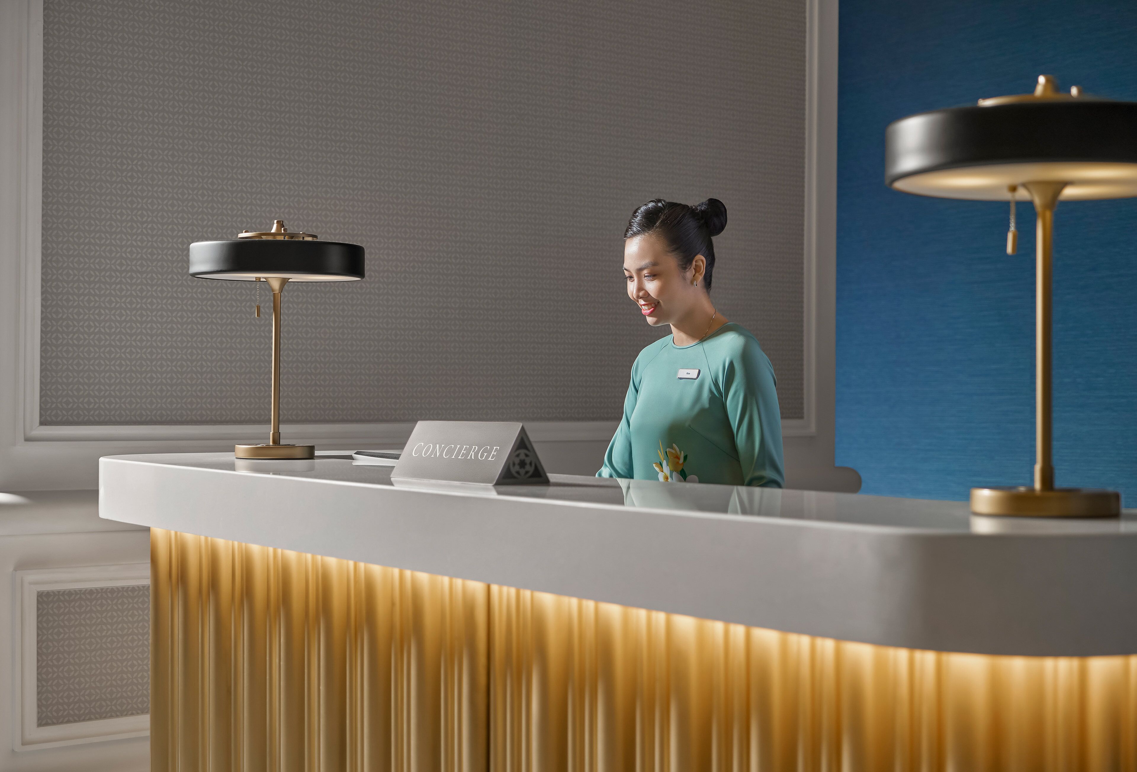 A reception desk with a nameplate labeled "CONCIERGE," a modern lamp, and a patterned wall in the background.