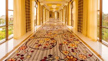 A long, well-lit corridor with large windows, a floral carpet, and a view of a pool and greenery outside.