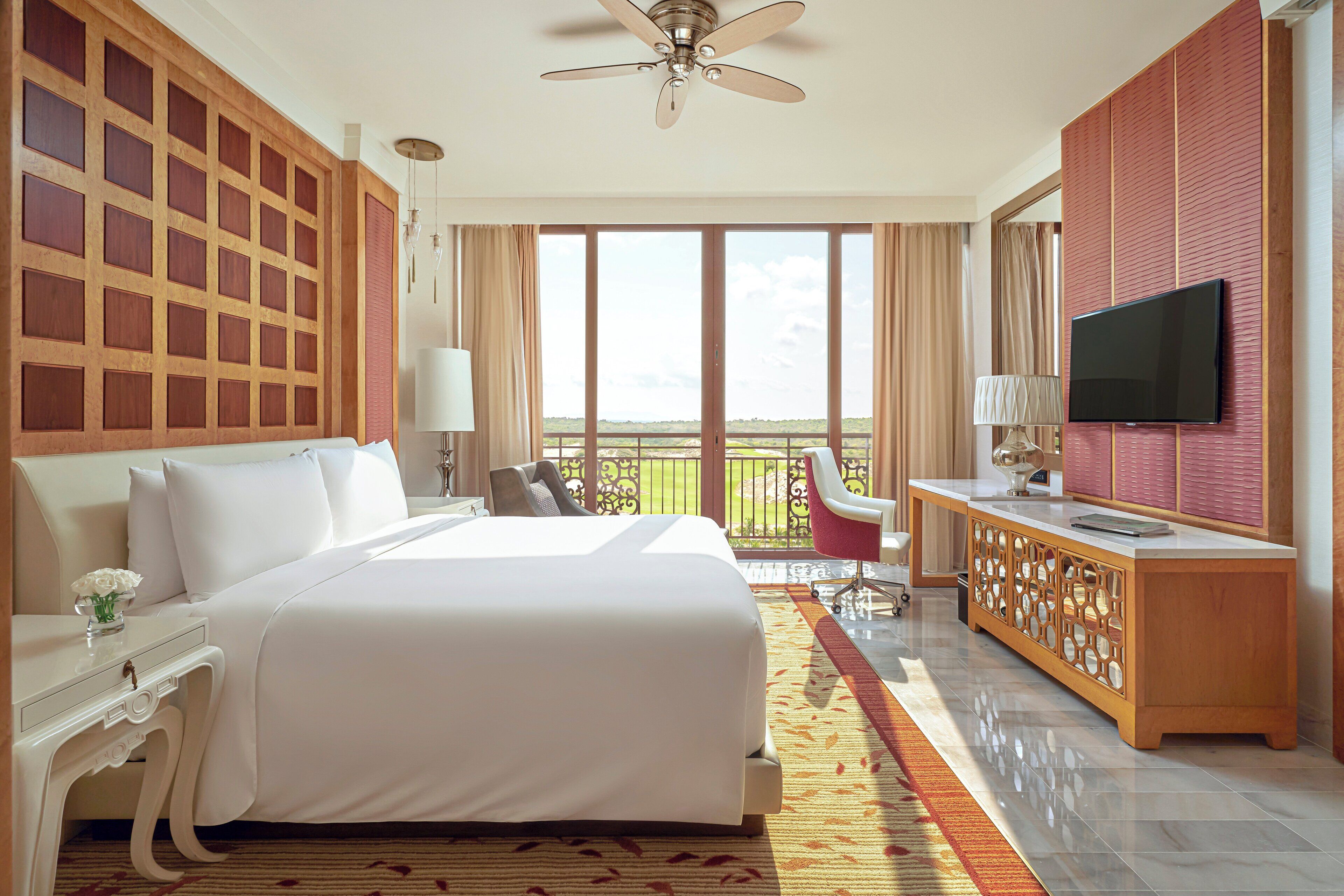 Quarto de hotel com uma cama grande, televisão, varanda com vista e ventoinha no teto.