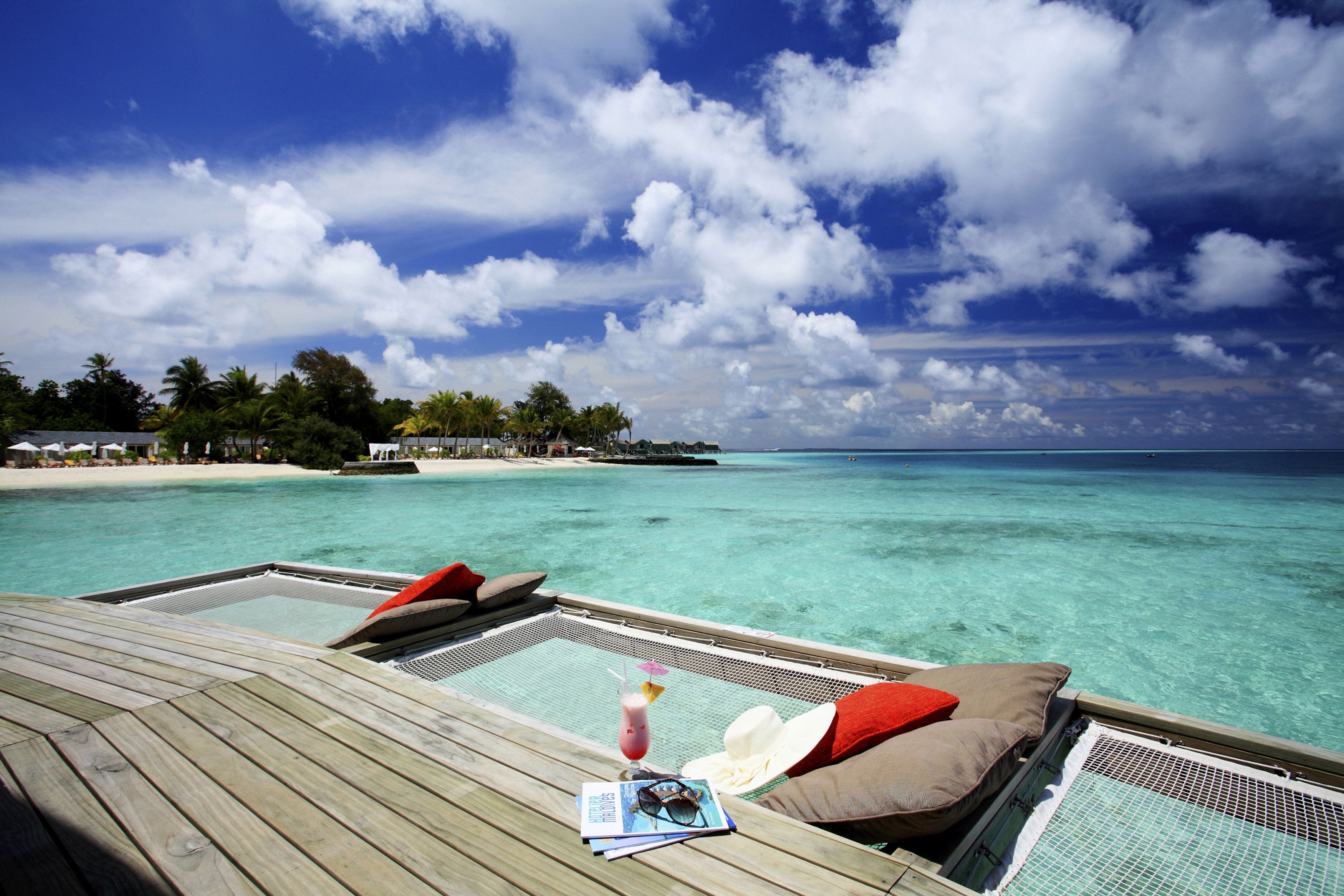 A wooden deck with lounge chairs and a drink, overlooking a clear turquoise sea and a beach with palm trees.