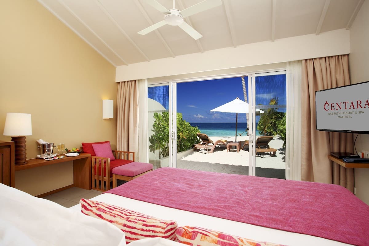 Habitación de hotel con cama, escritorio, silla y televisor. La habitación tiene vista a una playa y palmeras.