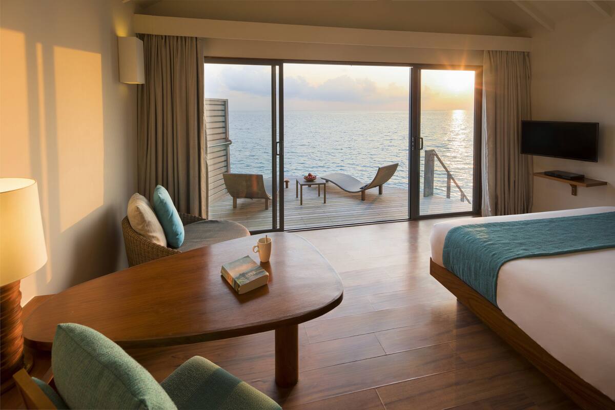 Habitación de hotel con cama, televisión, mesa con un libro y una taza, una silla y vista al mar a través de una puerta corredera.