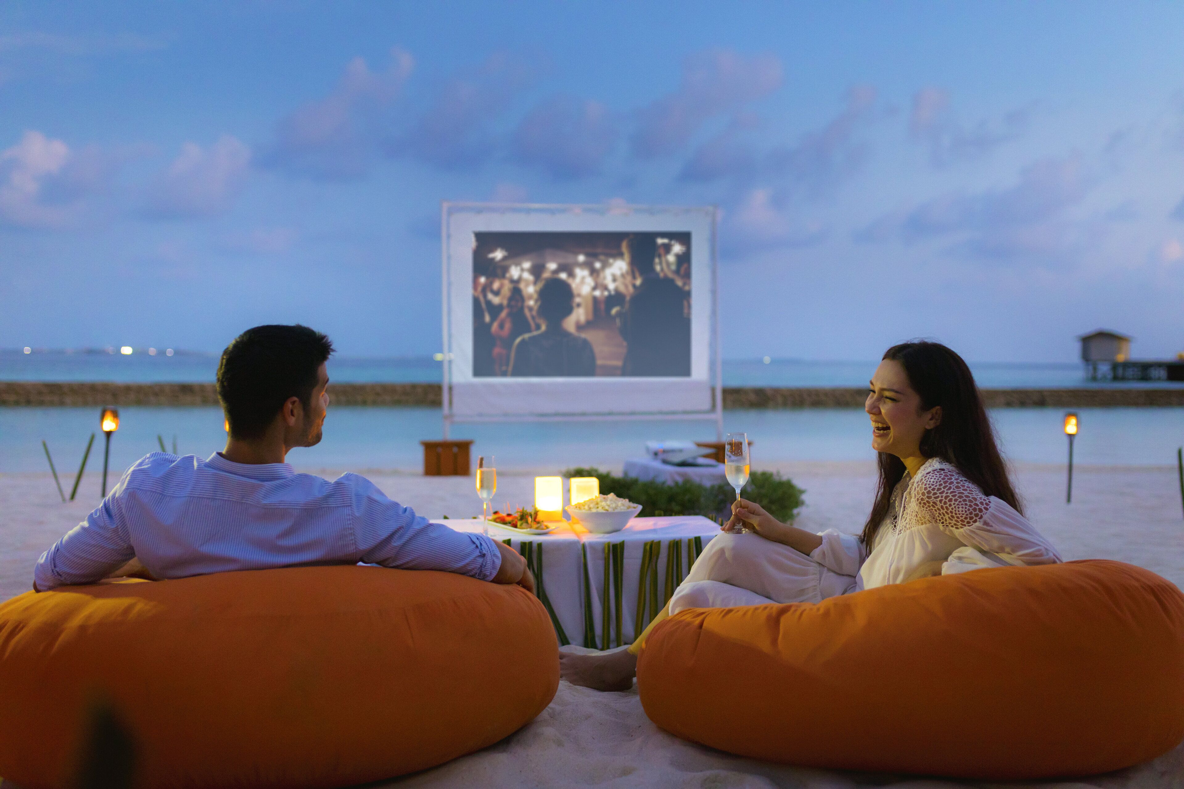 Dos personas sentadas en pufs mirando una mesa con una pantalla de proyector, comida y bebidas.