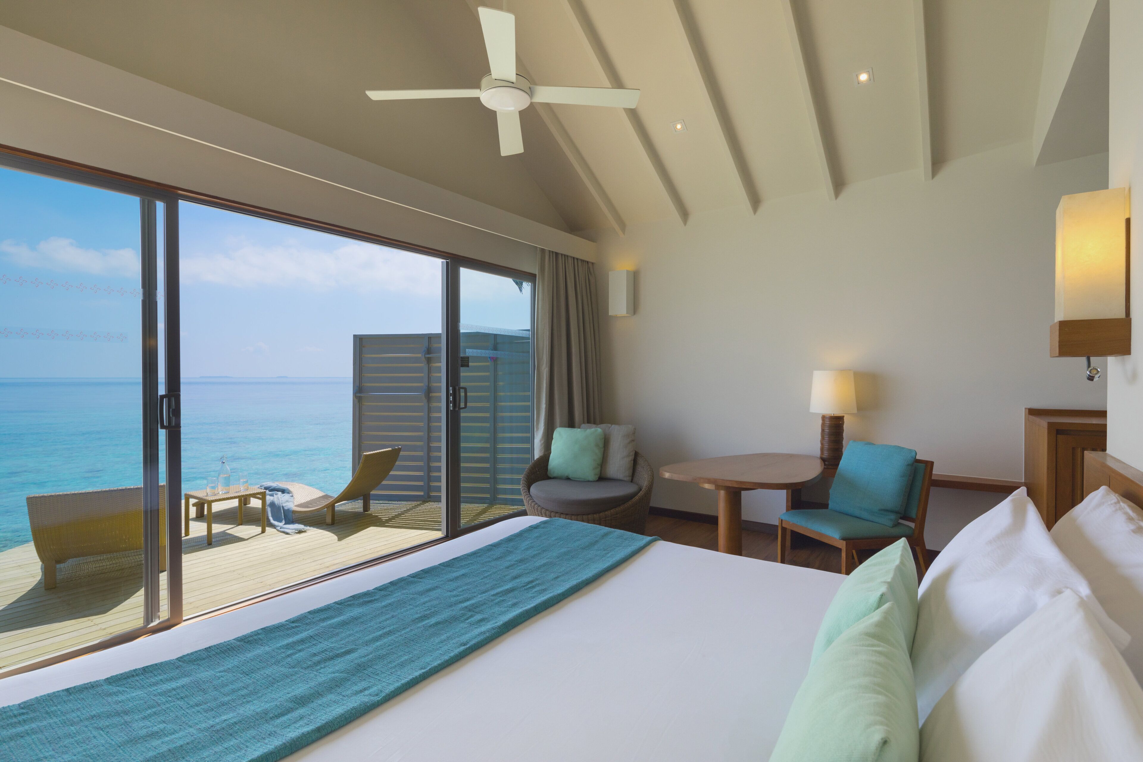 Quarto de hotel com uma cama grande, varanda com vista para o mar, ventilador de teto e uma pequena mesa com abajur.
