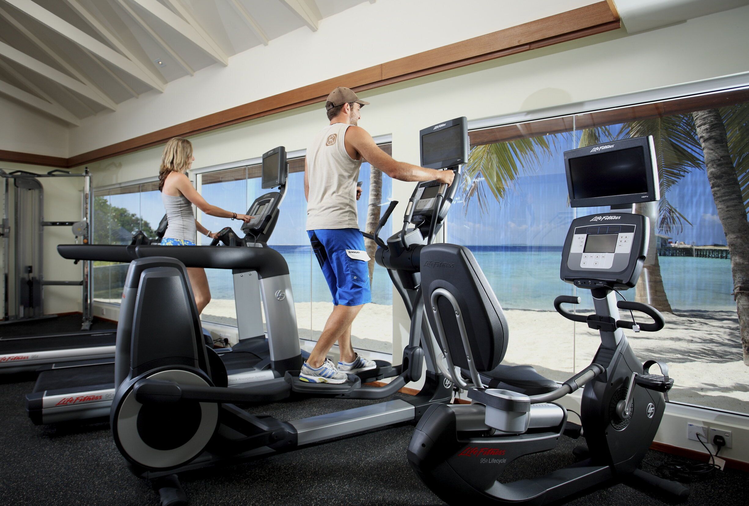 Une salle de sport équipée de tapis de course et de vélos d’exercice, avec de grandes fenêtres donnant sur la plage.