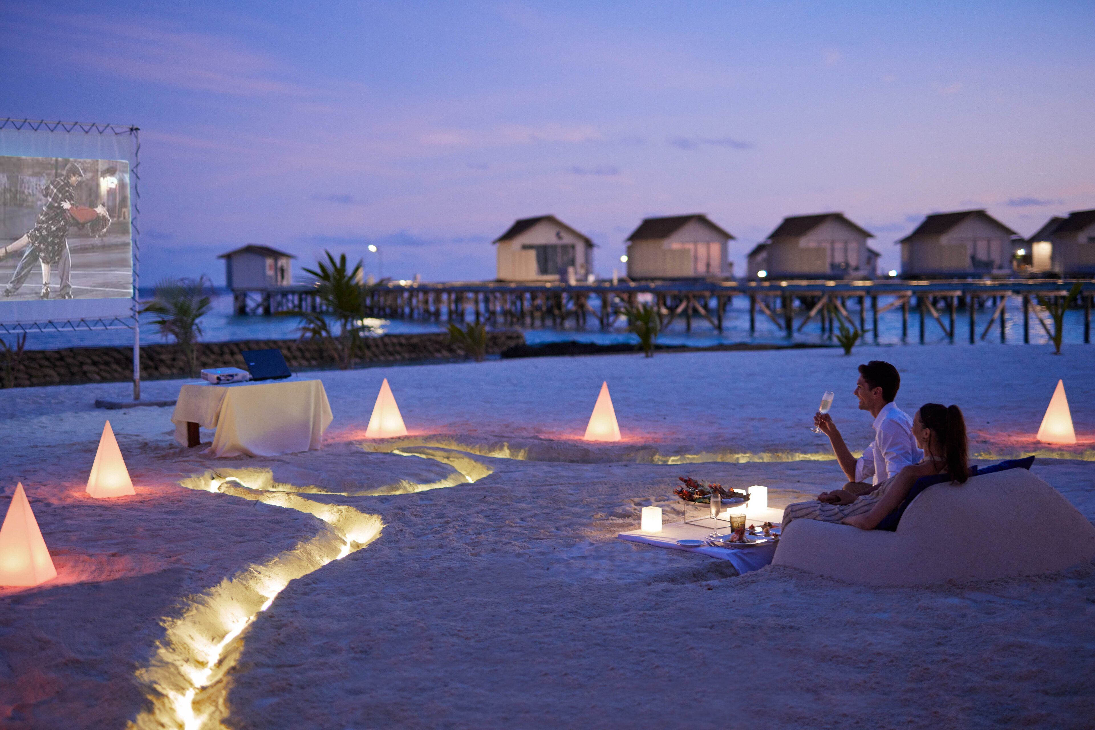 Un couple se détend sur une dune de sable, une table est dressée pour un repas, et ils ont une vue imprenable sur des bungalows sur pilotis et une jetée en bois.