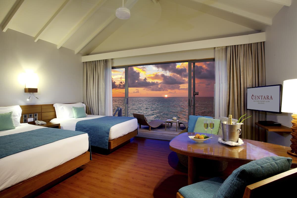 Habitación de hotel con dos camas, una mesa de comedor y vistas a la puesta de sol sobre el mar, con un cartel que dice "CENTARA".