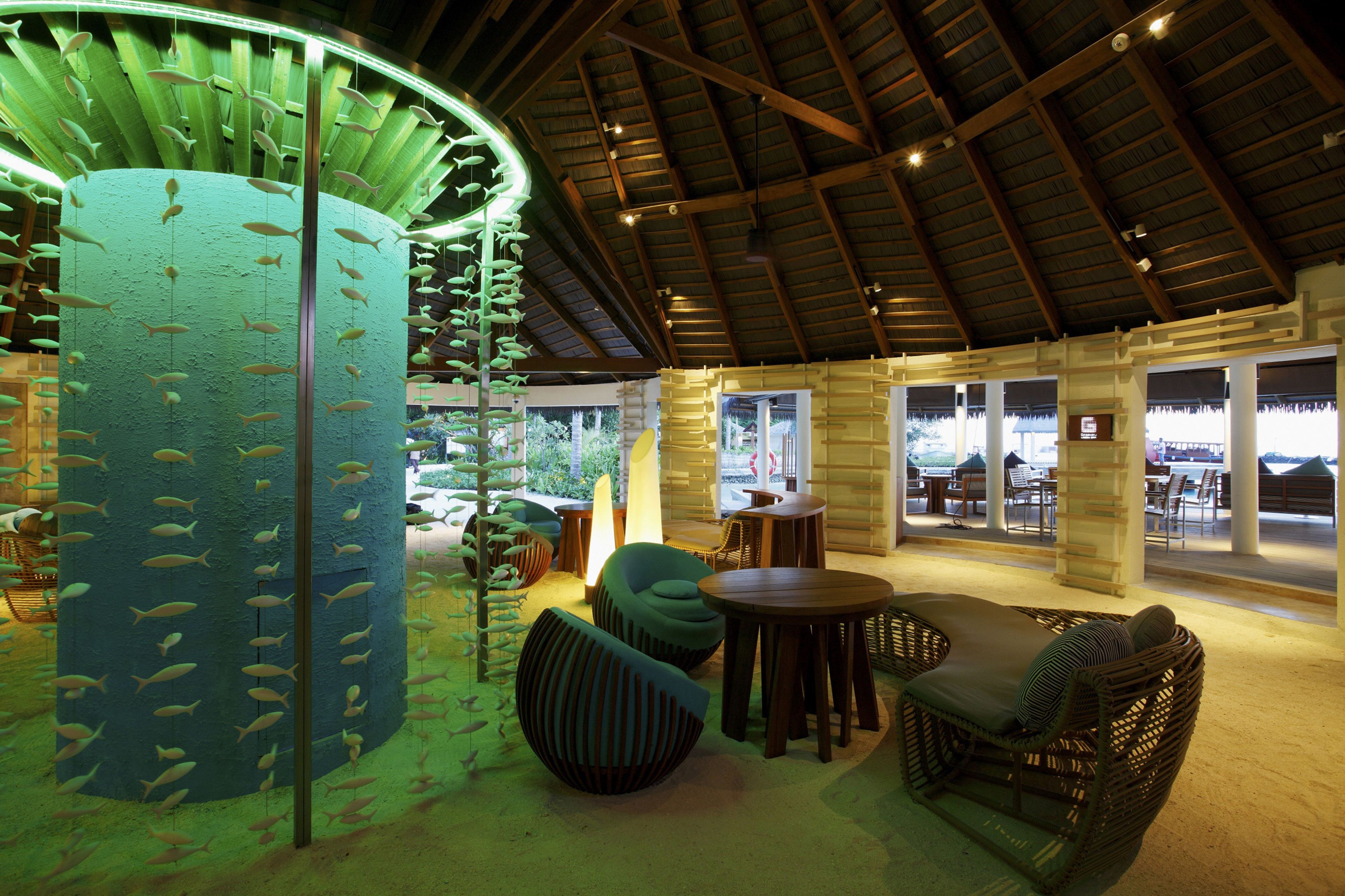 A spacious indoor area with a high ceiling, wooden beams, and a large cylindrical structure with hanging fish decorations. There are several seating areas with wicker furniture and a view of the outside through large windows.