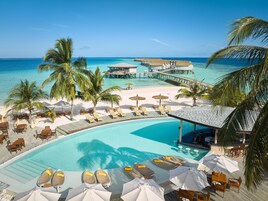 Zona de piscina en un resort con sillones de playa y sombrillas, vista despejada del océano y un muelle que se adentra en el agua.