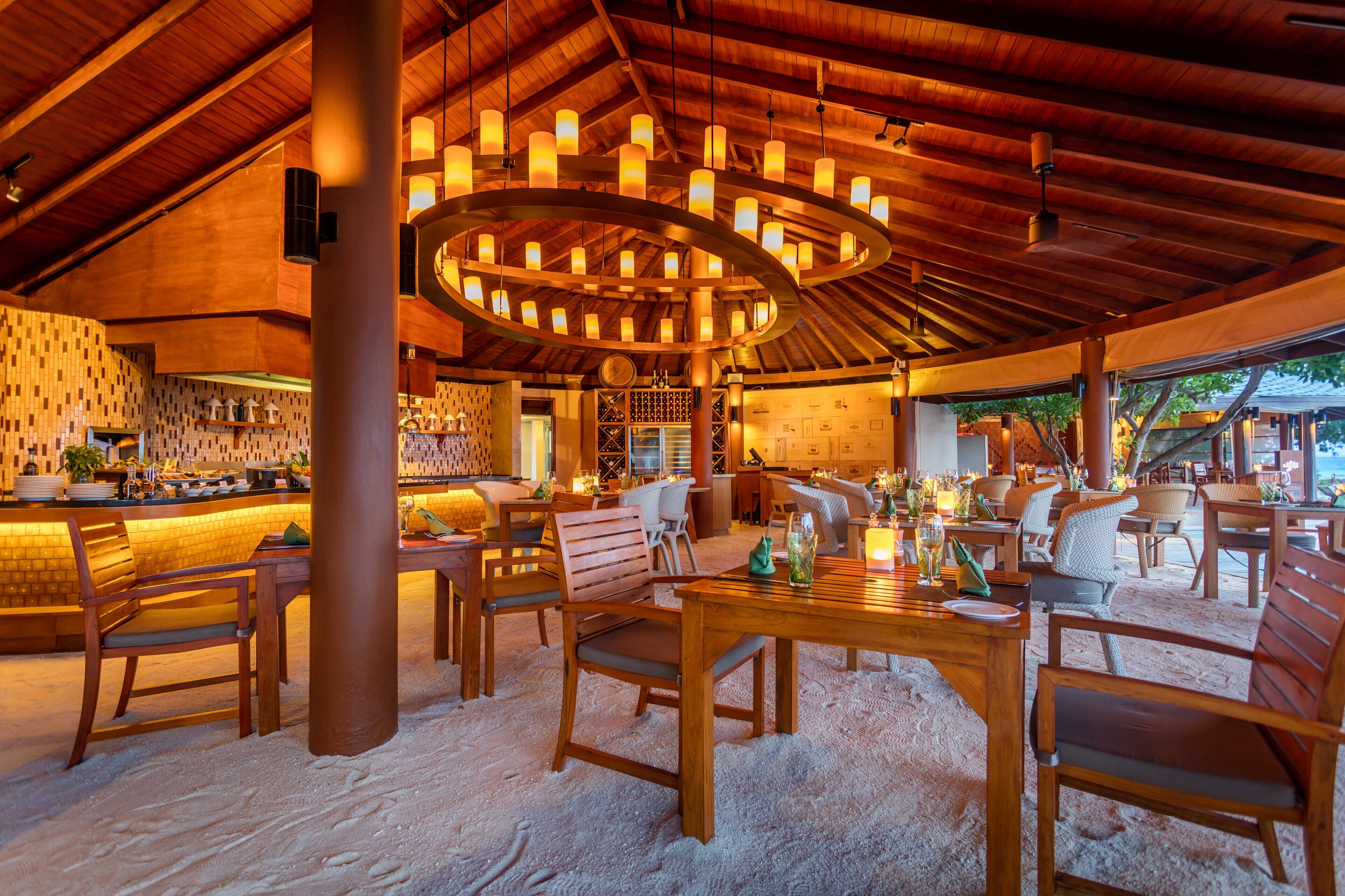 Un espacio interior amplio con techo alto y vigas de madera, que cuenta con una lámpara circular central, varias disposiciones de asientos y una zona de bar con salpicadero de azulejos.