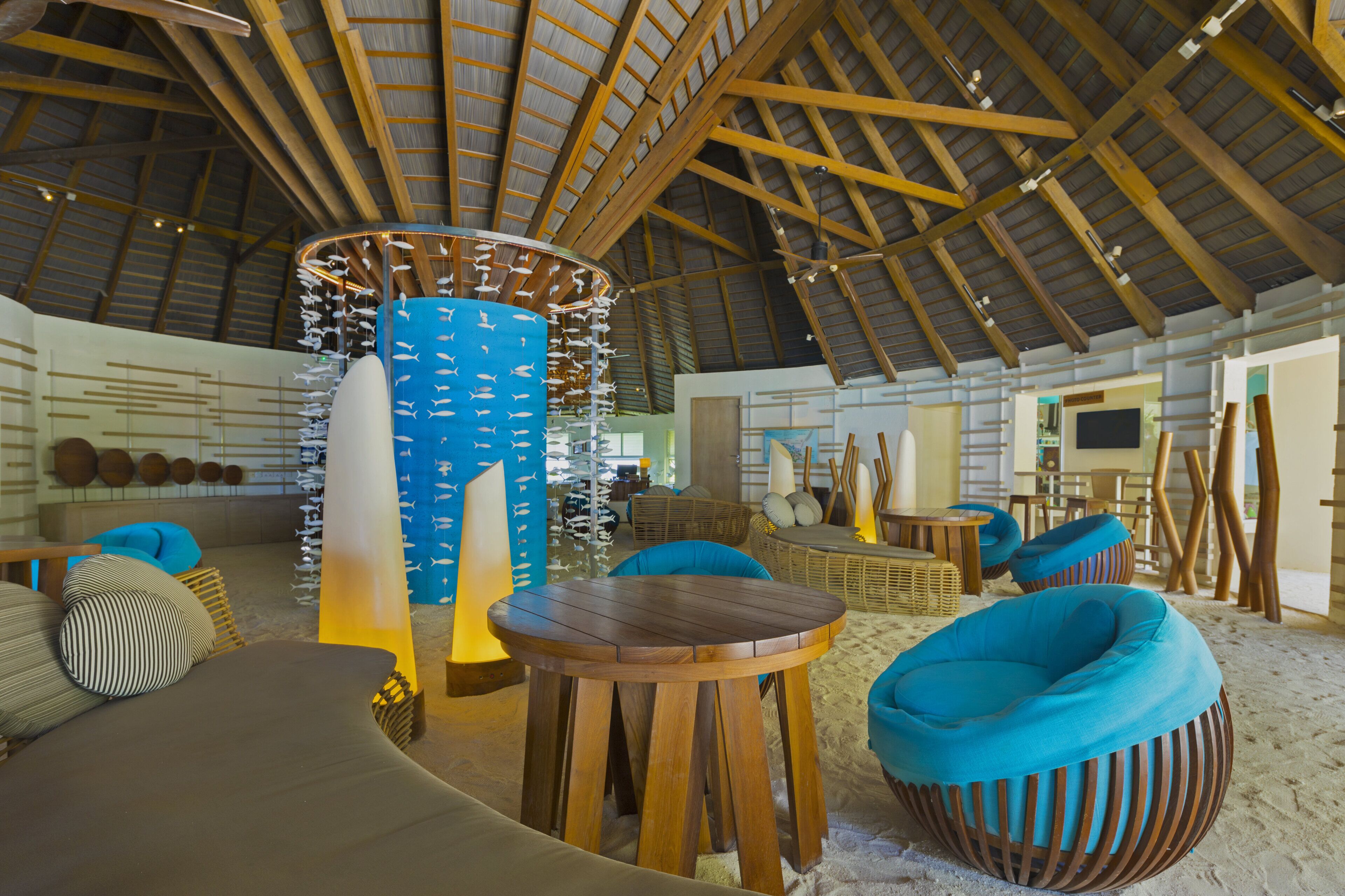 Une pièce spacieuse avec un haut plafond en bois, un pilier central orné de décorations suspendues, et plusieurs coins salon, dont une table ronde en bois et deux fauteuils bleus en forme de sac.