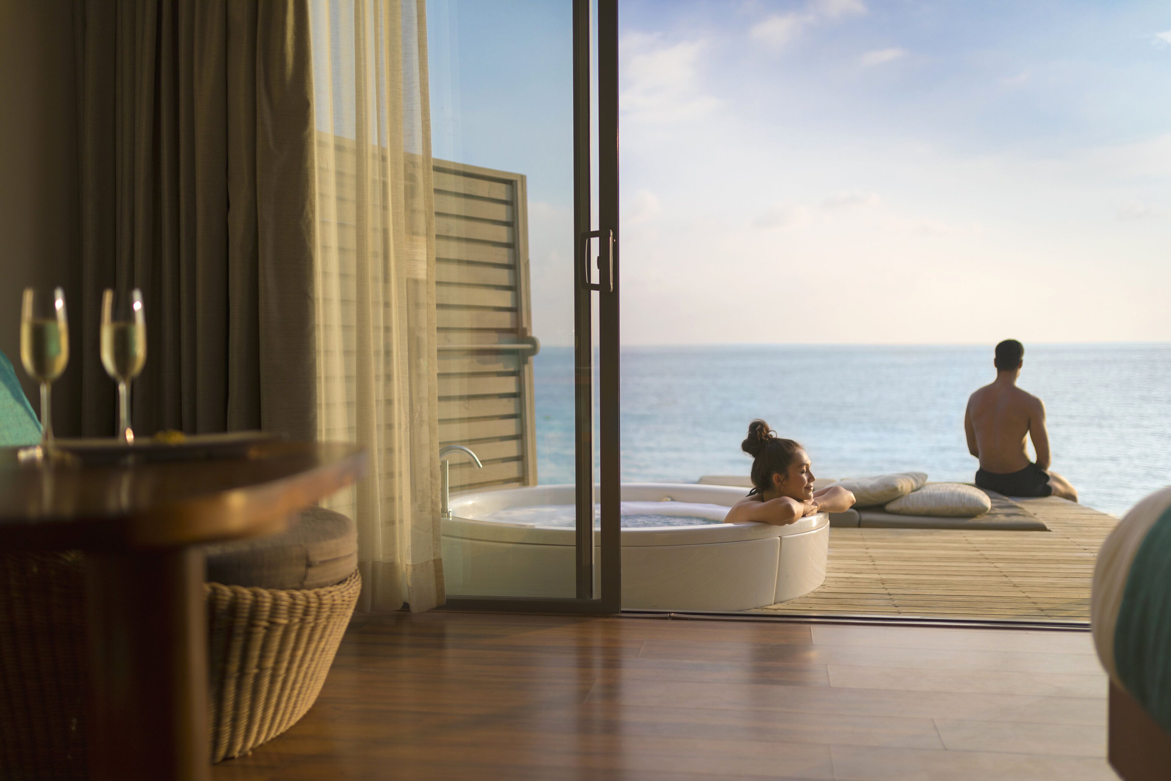 A room with a large window overlooking the sea, a bathtub, and a wooden deck.