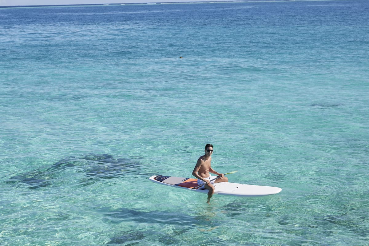 Una persona sentada en una tabla de surf en agua cristalina, otra persona relajándose en un jacuzzi, y vista al océano.