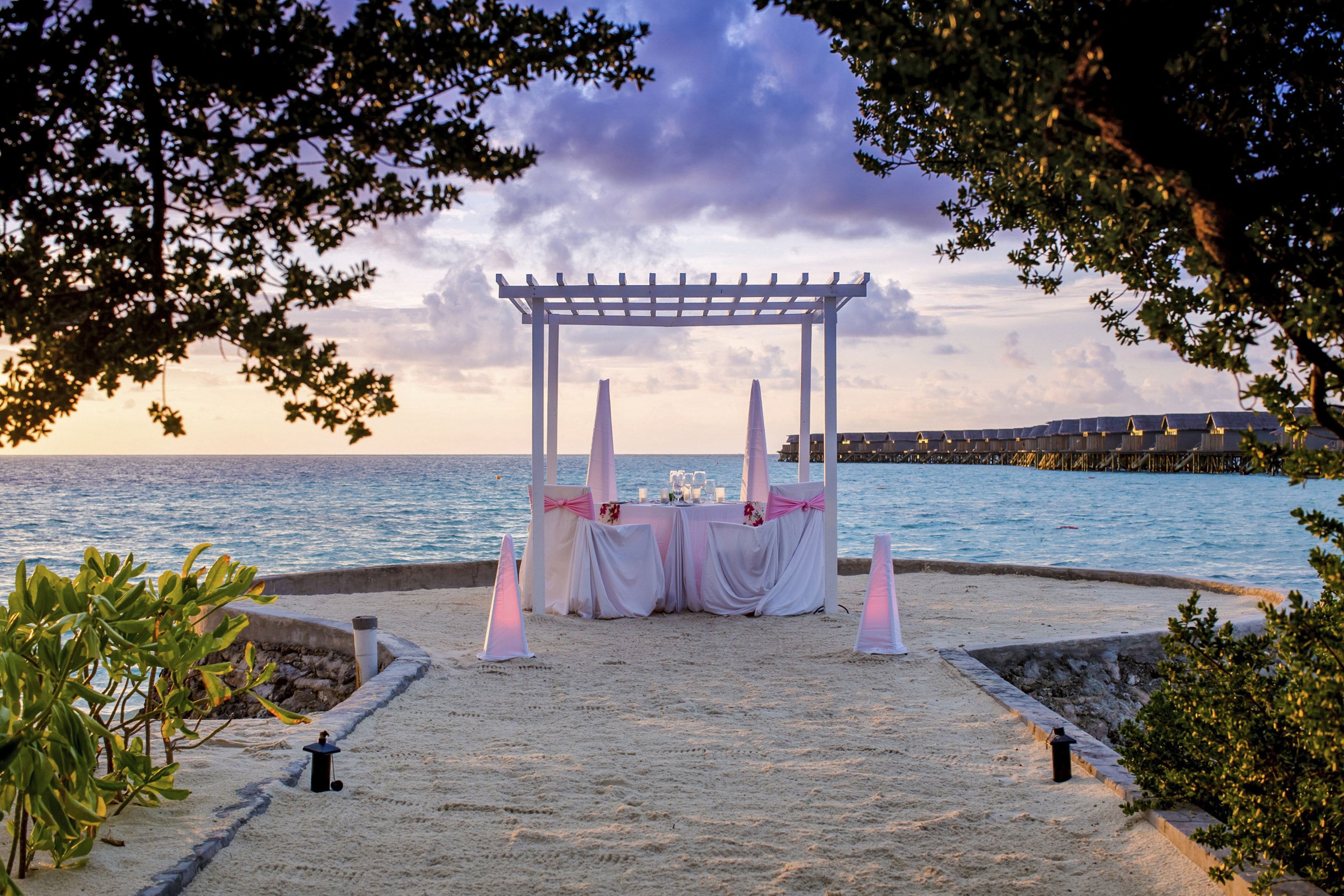 Een bruiloftdecoratie op het strand met een witte pergola, tafels met roze draperingen en kaarsen.