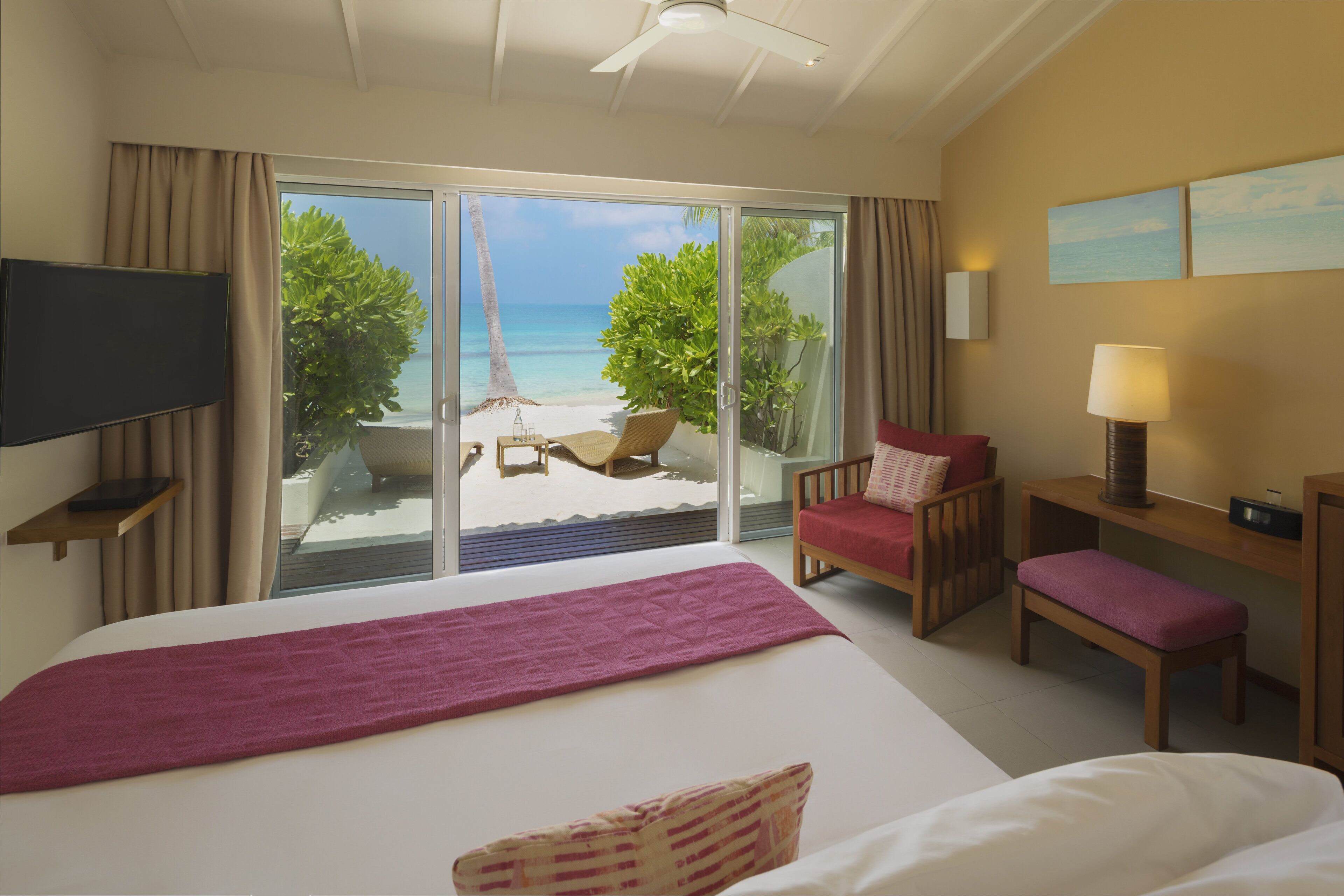 A hotel room with a large bed, a TV, a desk, a chair, a bench, and a view of a beach and palm trees.