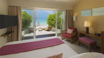 A hotel room with a large bed, a TV, a desk, a chair, a bench, and a view of a beach and palm trees.