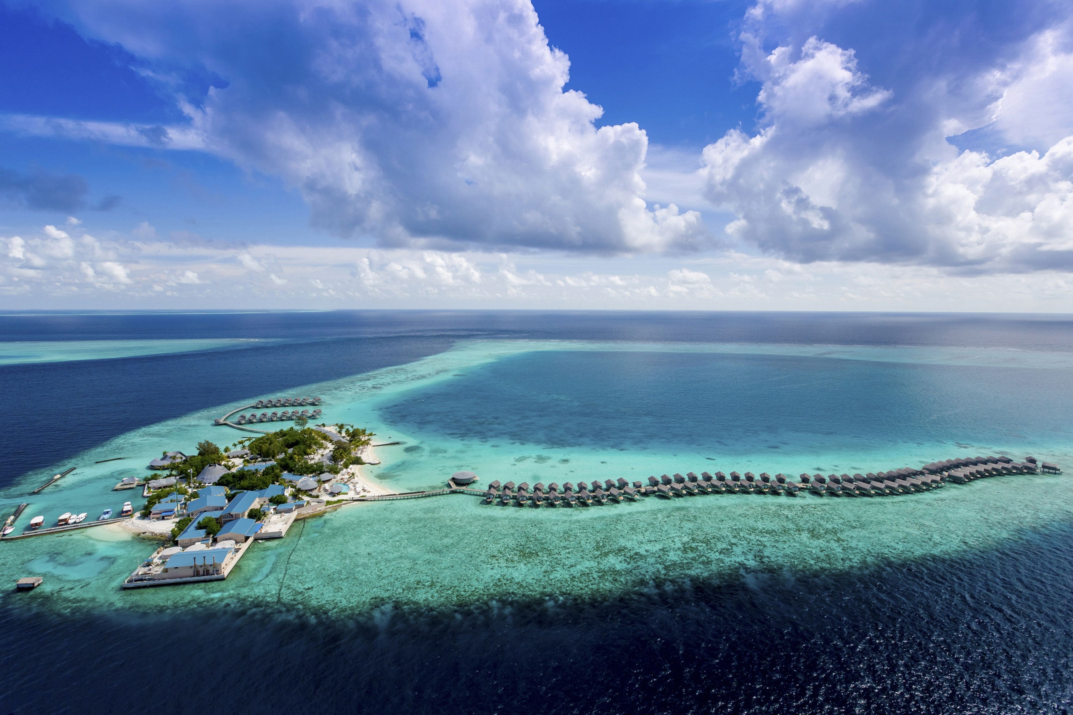 An aerial view of a tropical island resort with overwater bungalows, a pier, and clear turquoise waters.