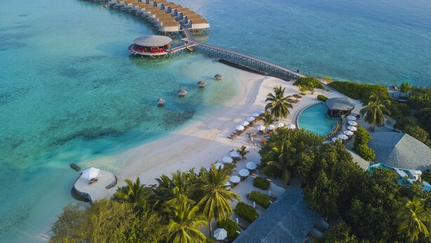 Un resort sobre el agua con un largo muelle que conecta con el edificio principal, aguas turquesas cristalinas y una zona de playa con sombrillas y tumbonas.