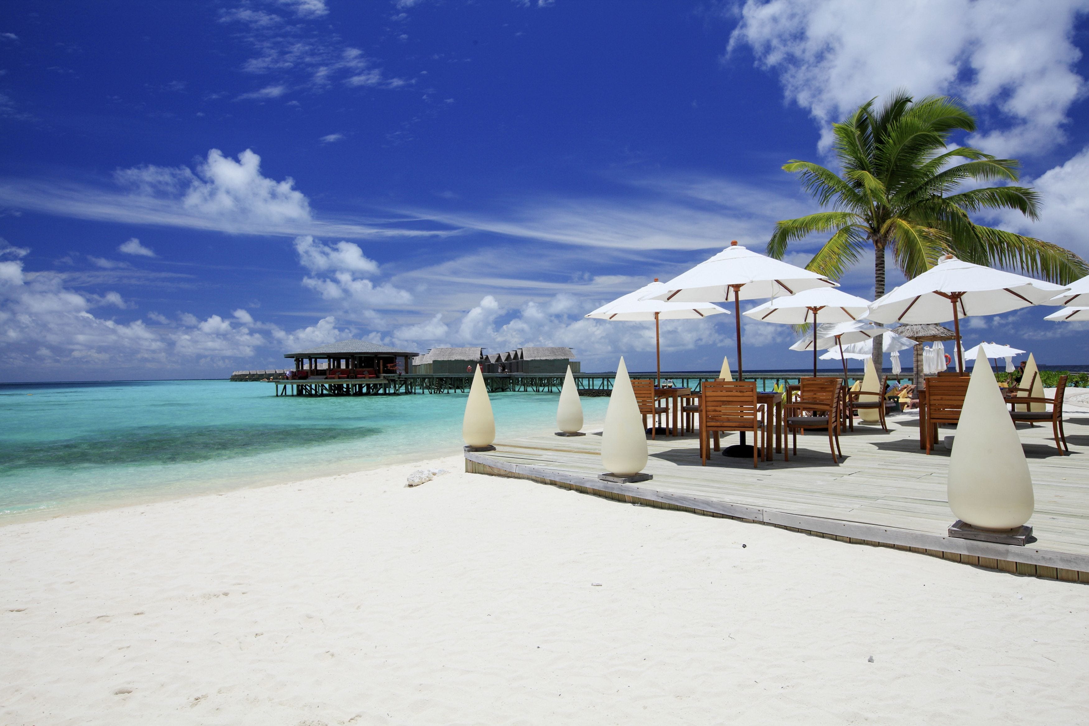 A beach resort with a wooden deck, white umbrellas, and a view of the ocean and overwater bungalows.