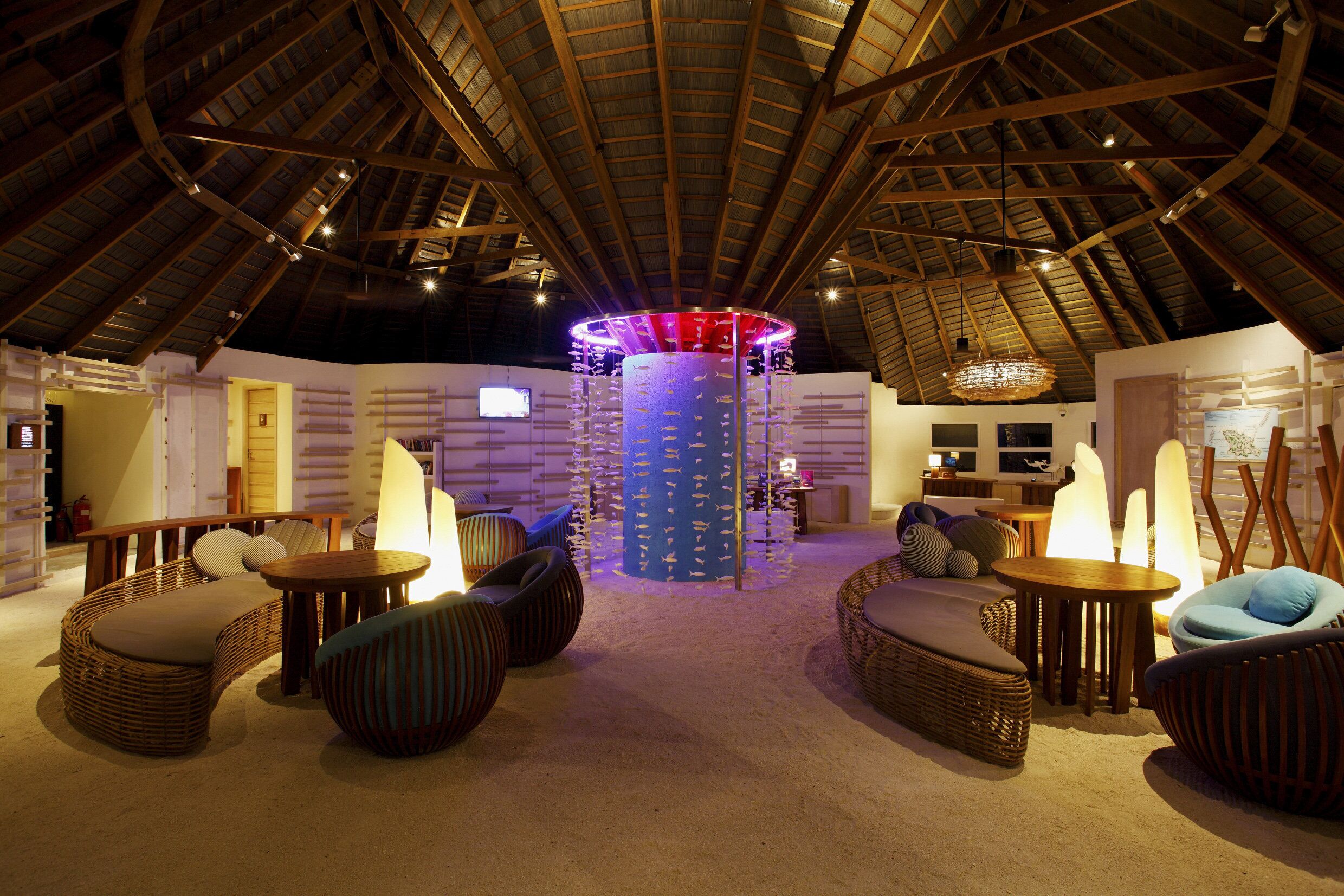 A spacious room with a high ceiling and a central column, furnished with wicker sofas and round tables, and a decorative light fixture.