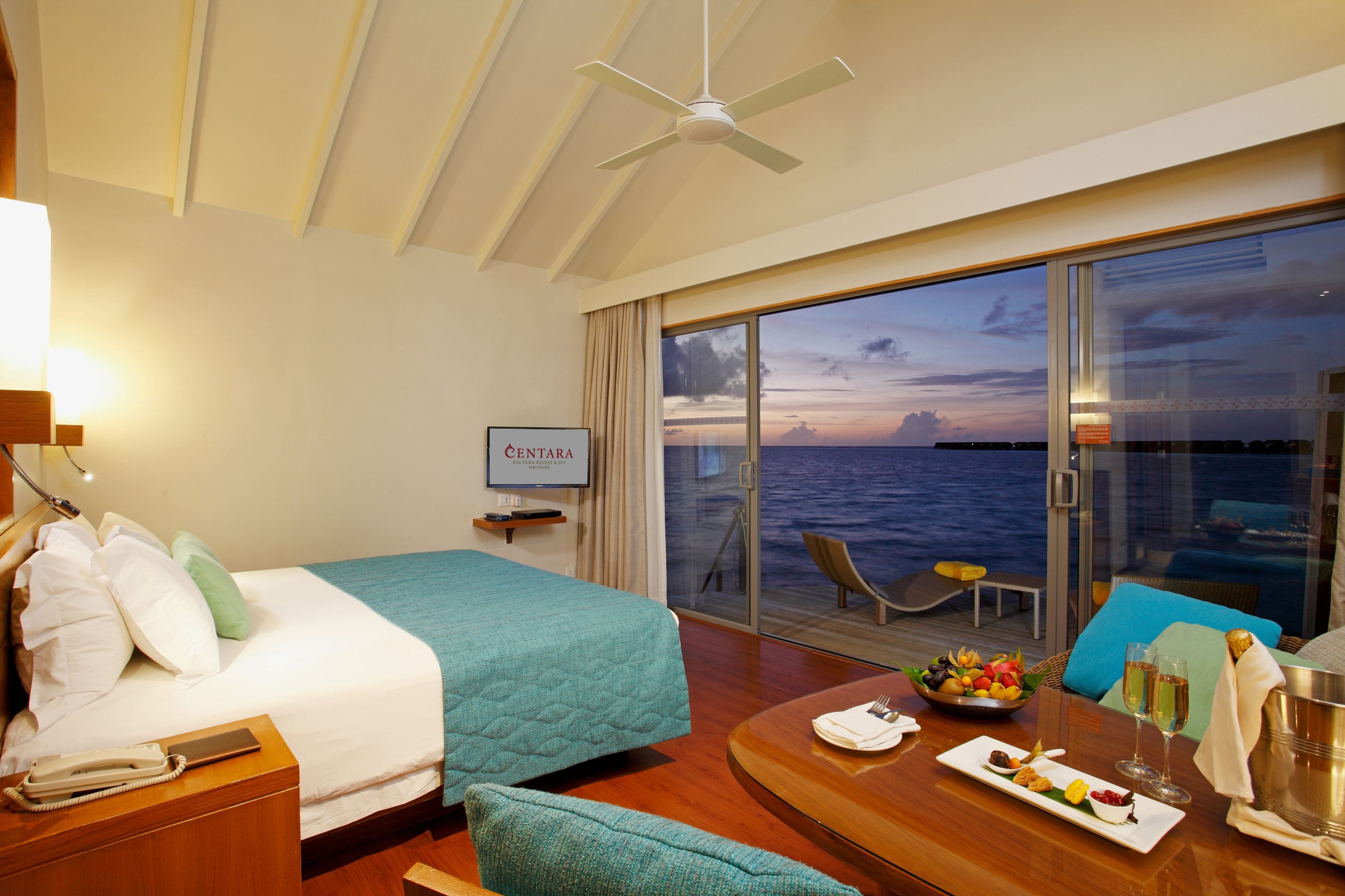 Ein Hotelzimmer mit Bett, einem Esstisch mit Obstteller und Gläsern, Blick auf das Meer durch große Fenster und einem Deckenventilator.