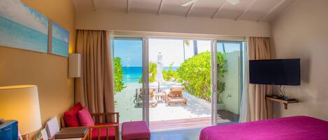 A hotel room with a bed, a desk with a chair, a TV, and a balcony with a view of the beach and ocean.
