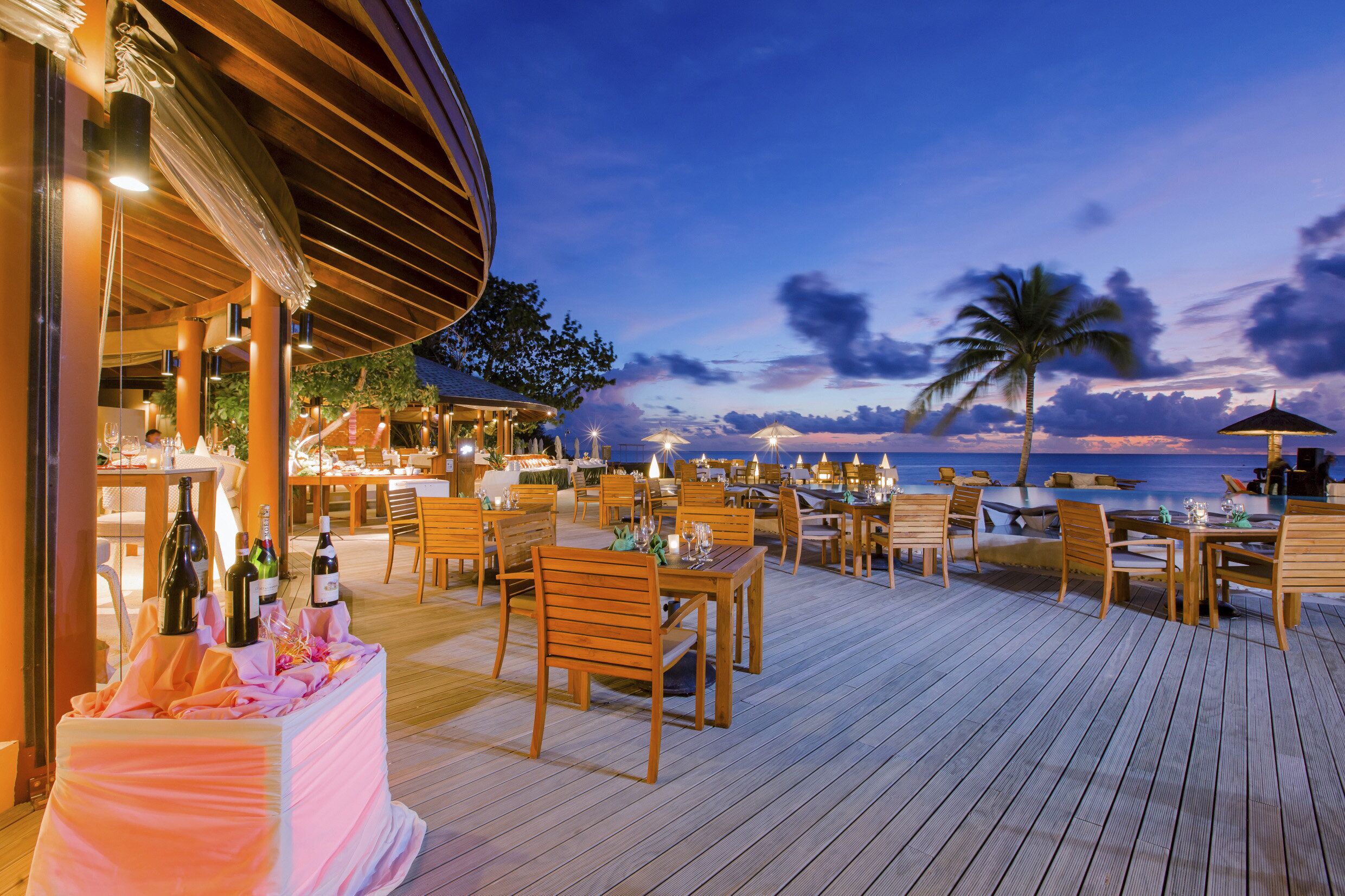 An outdoor dining area with wooden tables and chairs, a covered seating area, and a view of the ocean at dusk.
