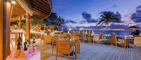 An outdoor dining area with wooden tables and chairs, a covered seating area, and a view of the ocean at dusk.