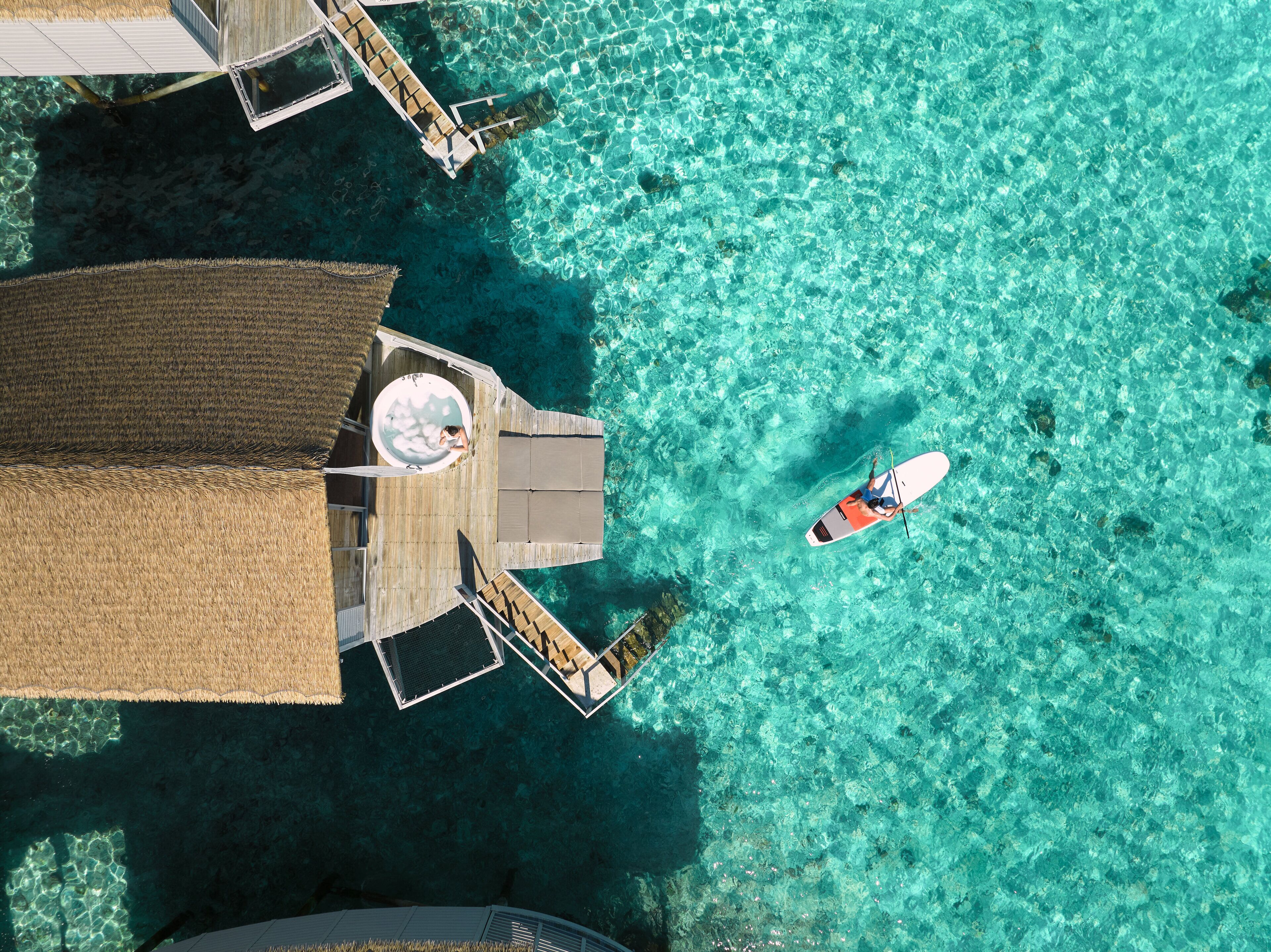 An aerial view of a resort with clear turquoise waters, a wooden deck, and a person paddleboarding.