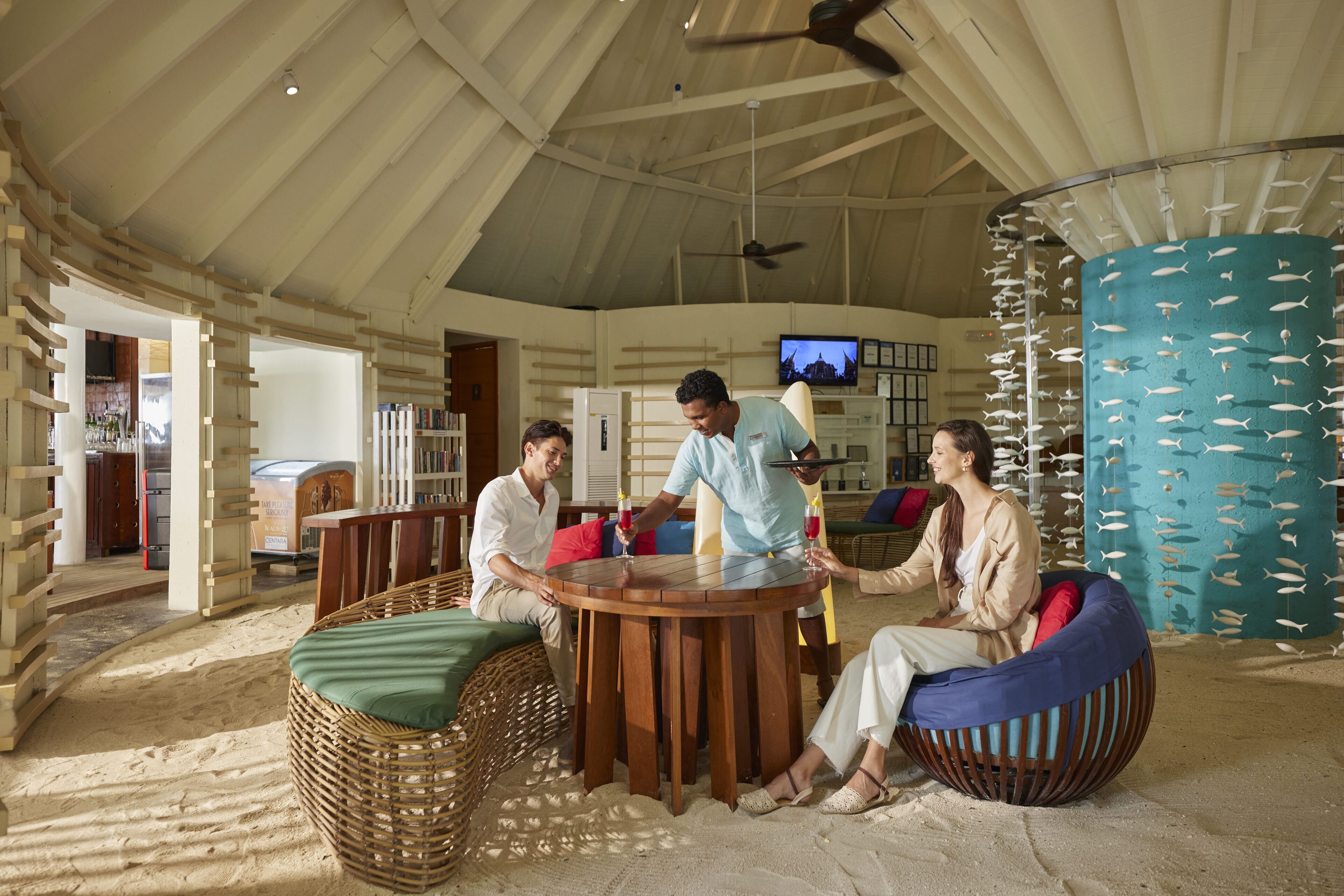 A group of people around a wooden table in a room with a high ceiling, a television, and a wall decorated with fish.