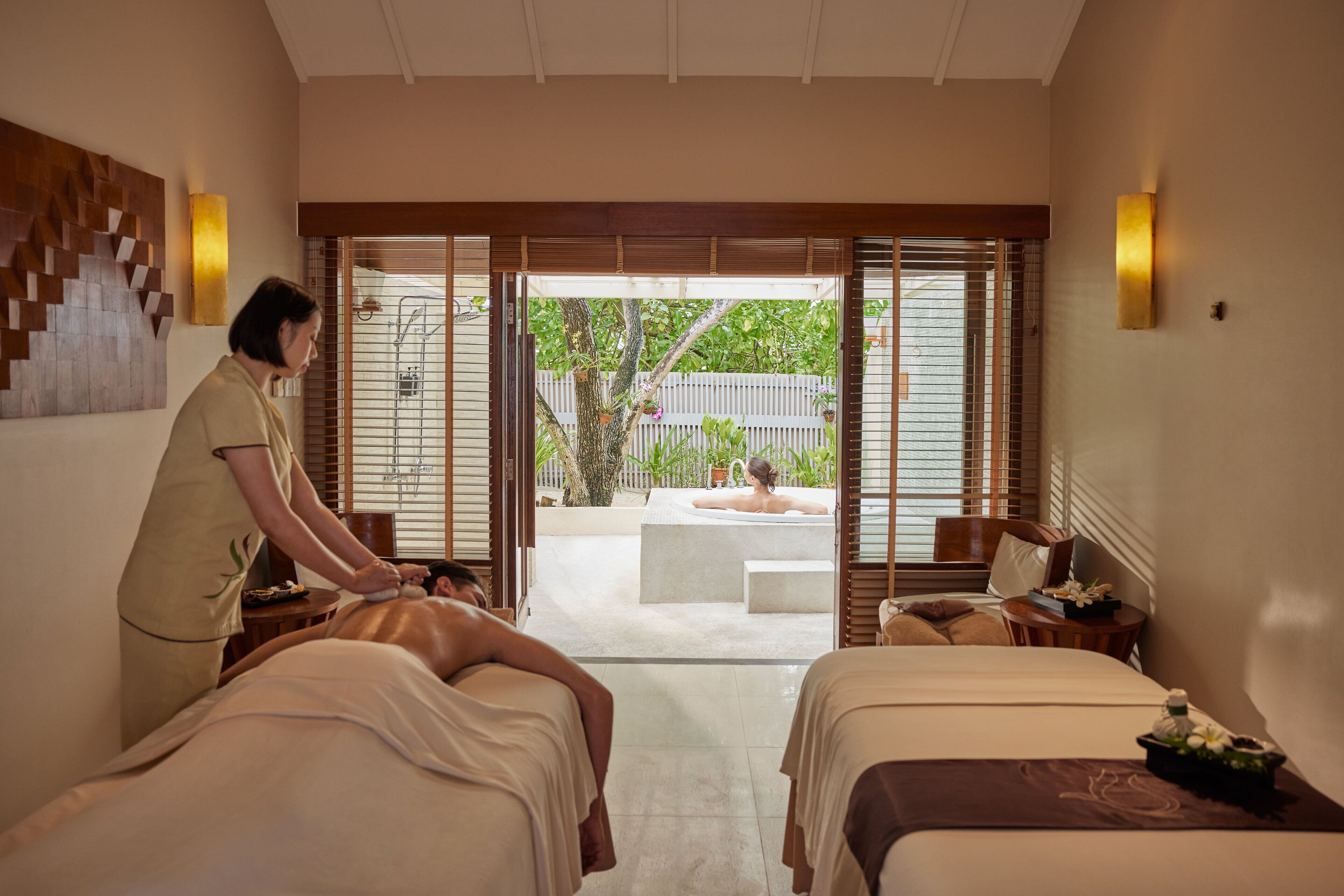 Een spa met twee massagetafels, een massagetherapeut en uitzicht op een tuin met een jacuzzi.