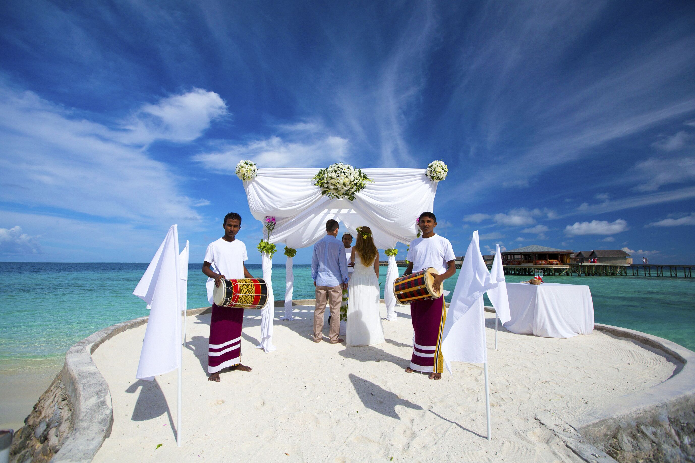 Ceremonia en la playa con un telón blanco drapeado y arreglos florales, con un cielo azul claro y el océano de fondo.