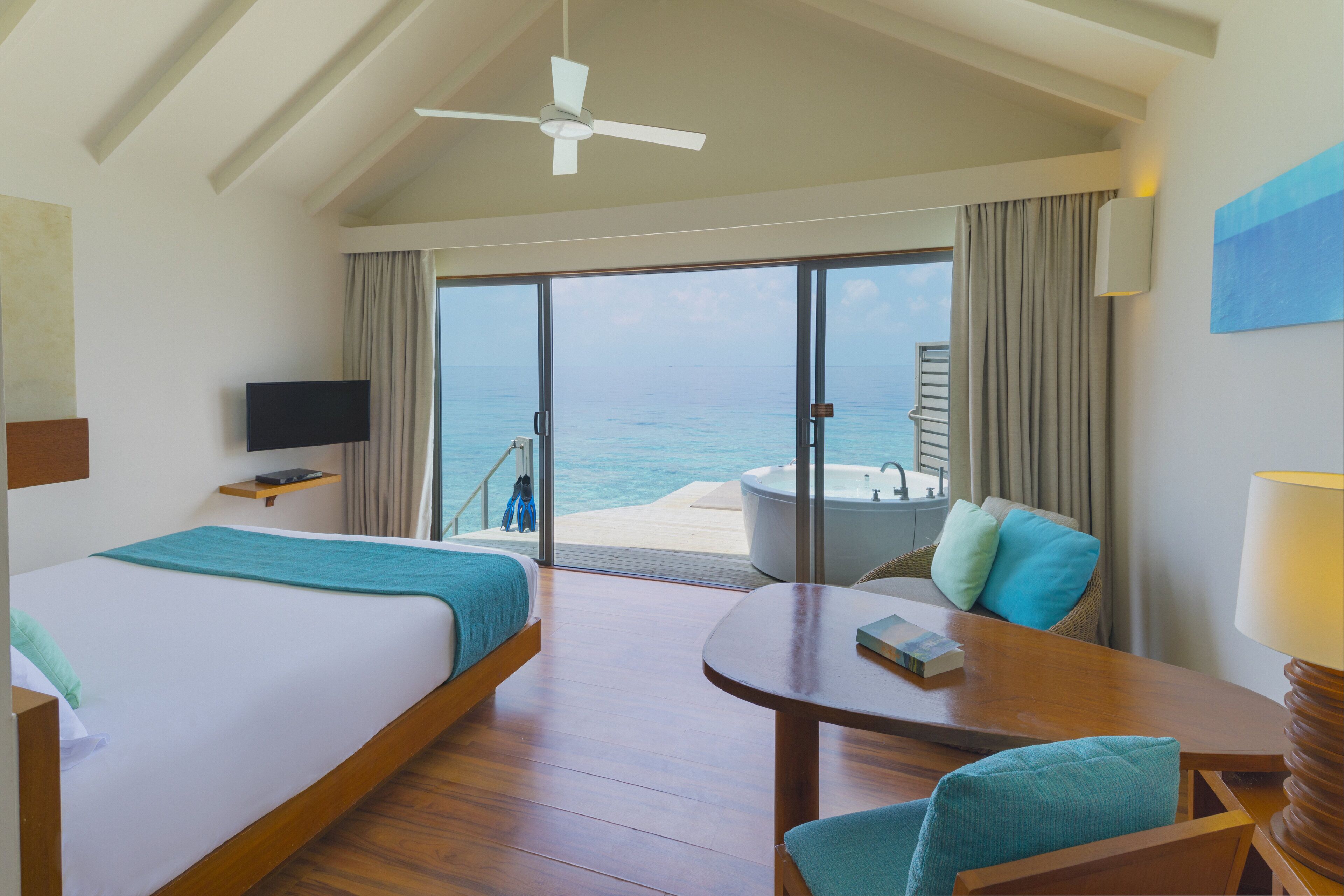 Habitación de hotel con dos camas, televisor, mesa, silla, lámpara, balcón con vista al mar y ventilador de techo.