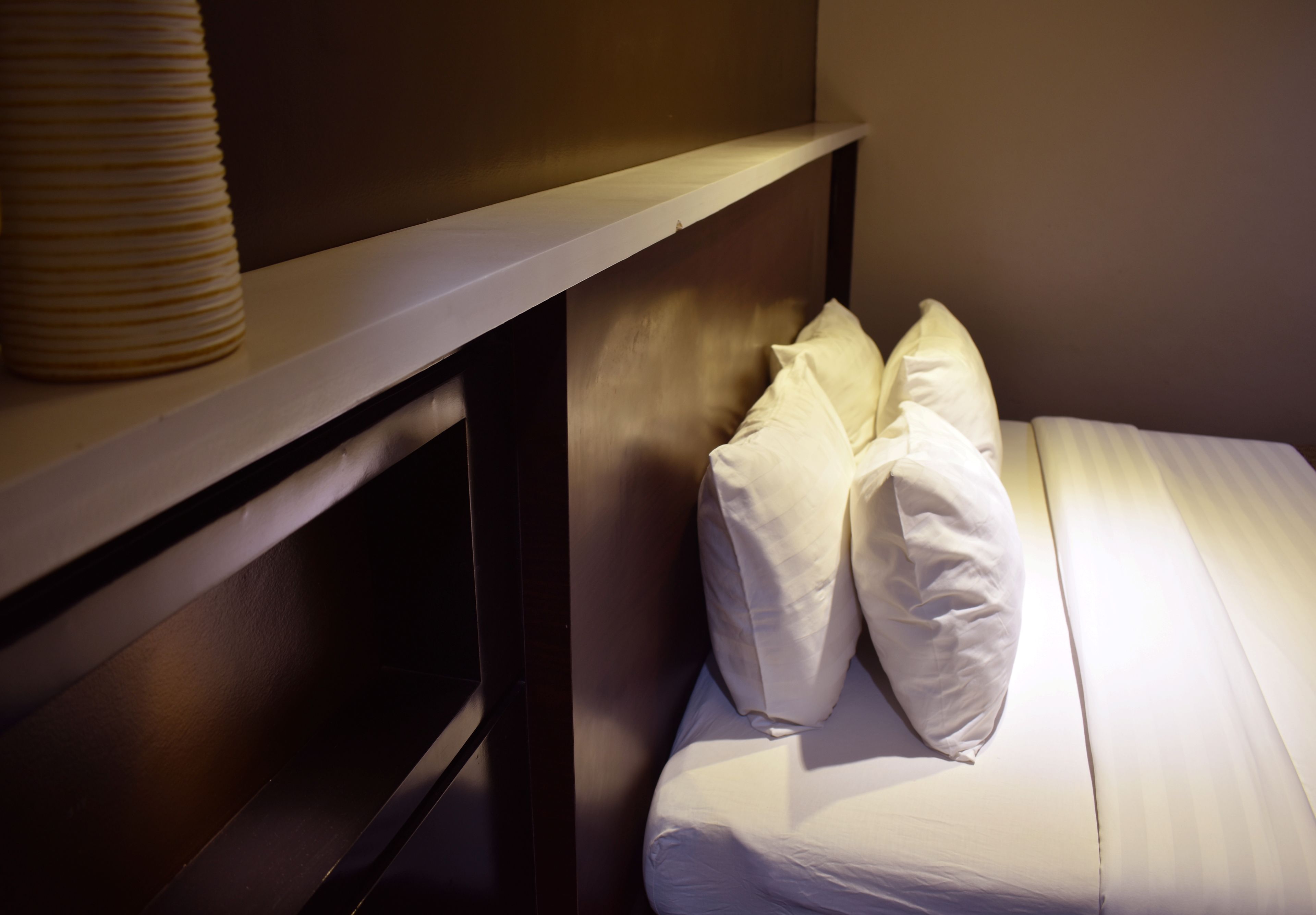A neatly made bed with white linens and pillows, a wooden headboard, and a shelf with a cylindrical container.
