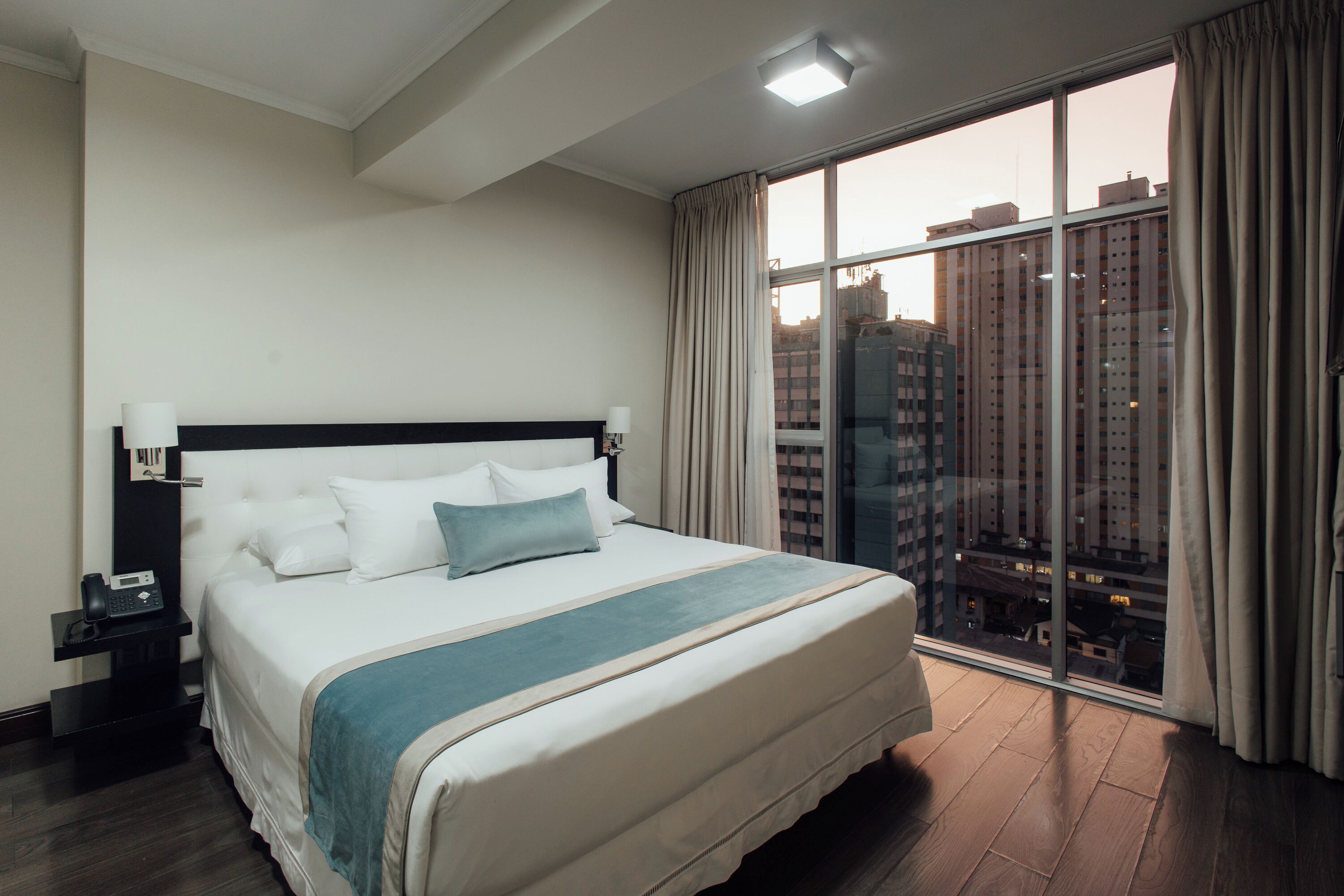 Habitación de hotel con una cama grande, una mesita de noche, un teléfono y vistas de edificios altos a través de un ventanal.