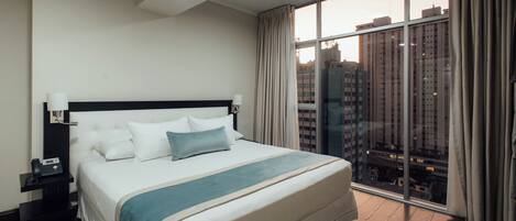 Habitación de hotel con una cama grande, una mesita de noche, un teléfono y vistas de edificios altos a través de un ventanal.