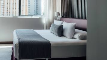 A neatly made bed with a striped blanket, a bedside lamp, and a view of tall buildings through a window.