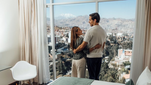Un couple se tient près d’une fenêtre, d’où l’on aperçoit un paysage urbain dominé par des montagnes.