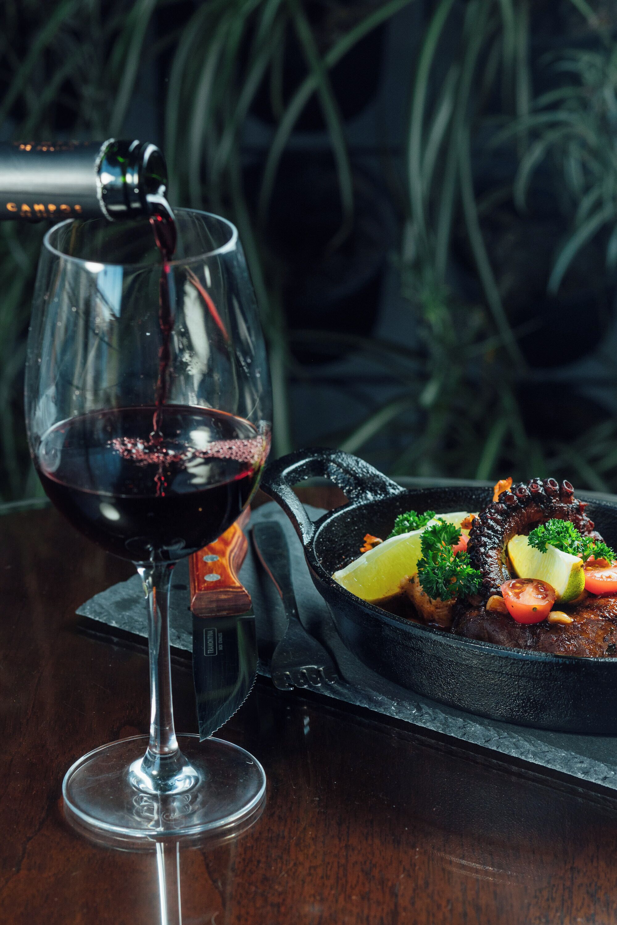 Una copa de vino tinto siendo servida, una sartén con comida, y un fondo oscuro con plantas.