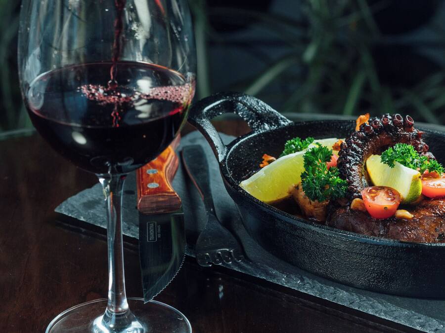 Una copa de vino tinto siendo servida, una sartén con comida, y un fondo oscuro con plantas.