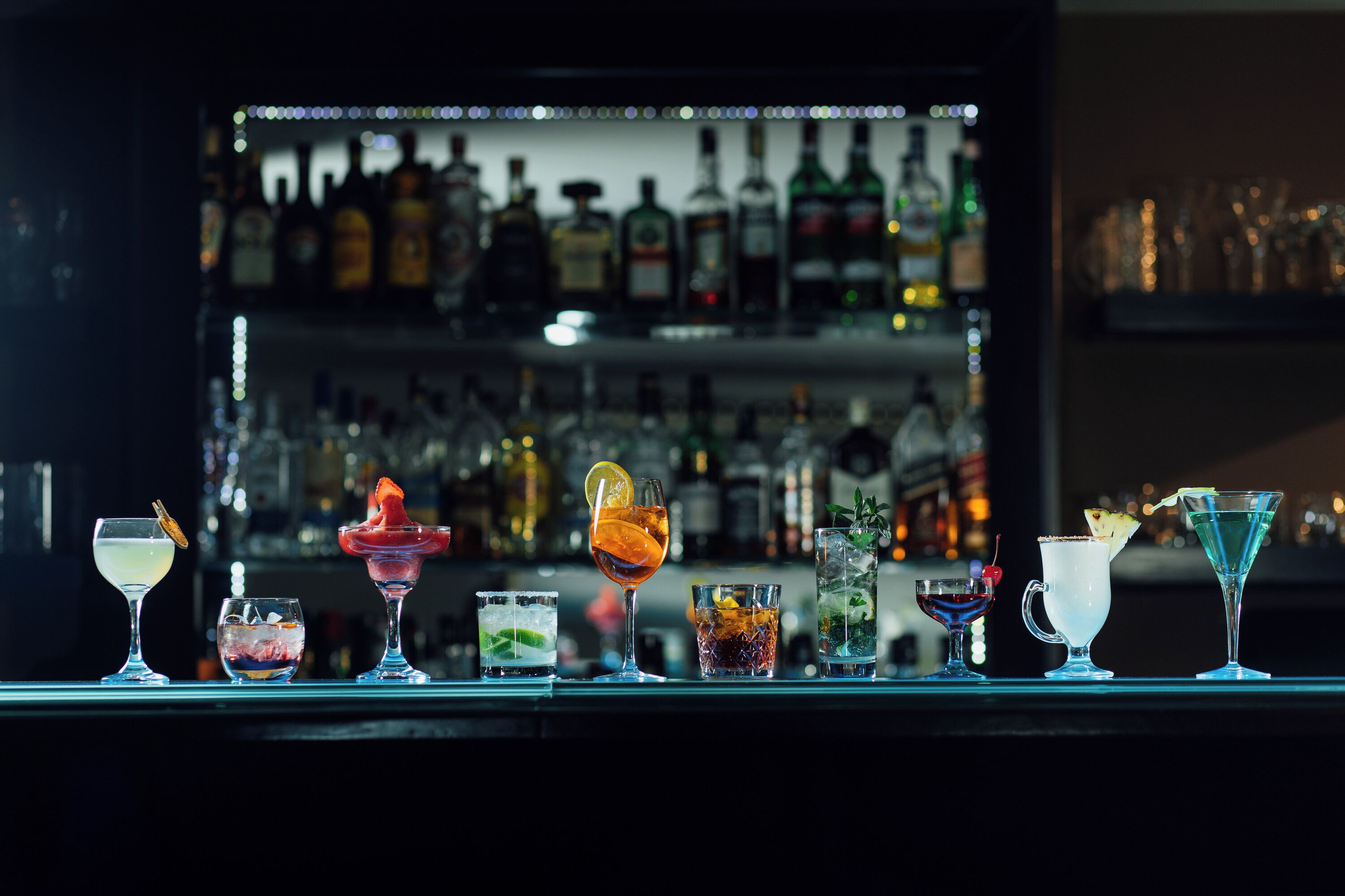 A bar counter with various cocktails and a background of shelves stocked with bottles.
