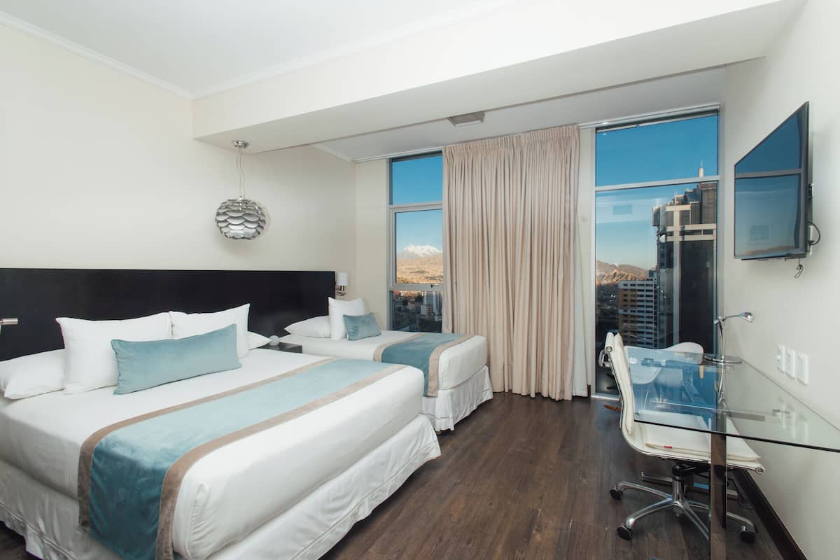 Habitación de hotel con dos camas, un escritorio de cristal, televisión y vistas a la ciudad.