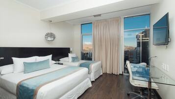 Quarto de hotel com duas camas, escrivaninha de vidro, televisão e vista para a cidade.