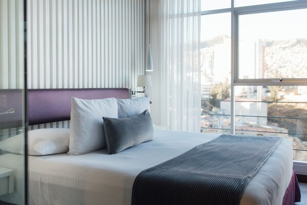 Habitación de hotel con cama, ventana con vista a edificios y mesita de noche con lámpara.